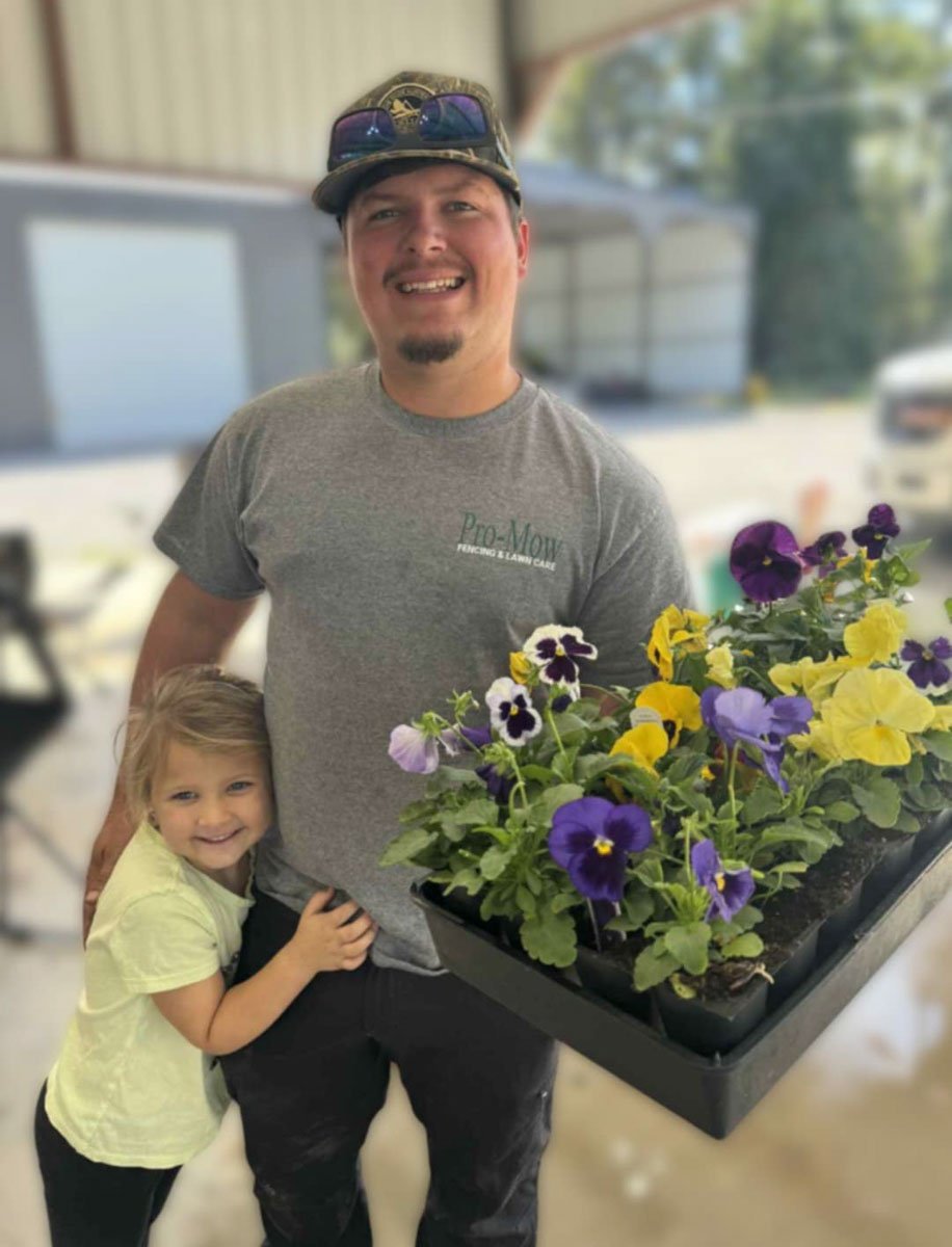 A man who works at Pro-Mow lawn care in Alexandria, LA is holding a tray of colorful pansies. A a young girl beside him is hugging his leg, both smiling for the picture.