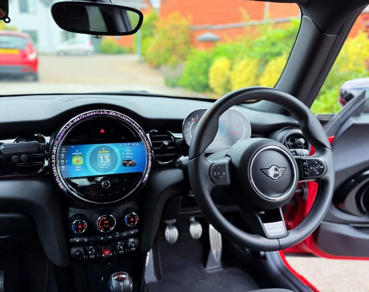 Interior of a Mini Cooper with a digital dashboard, steering wheel, and outdoor view through the windshield showing cars and colorful bushes.