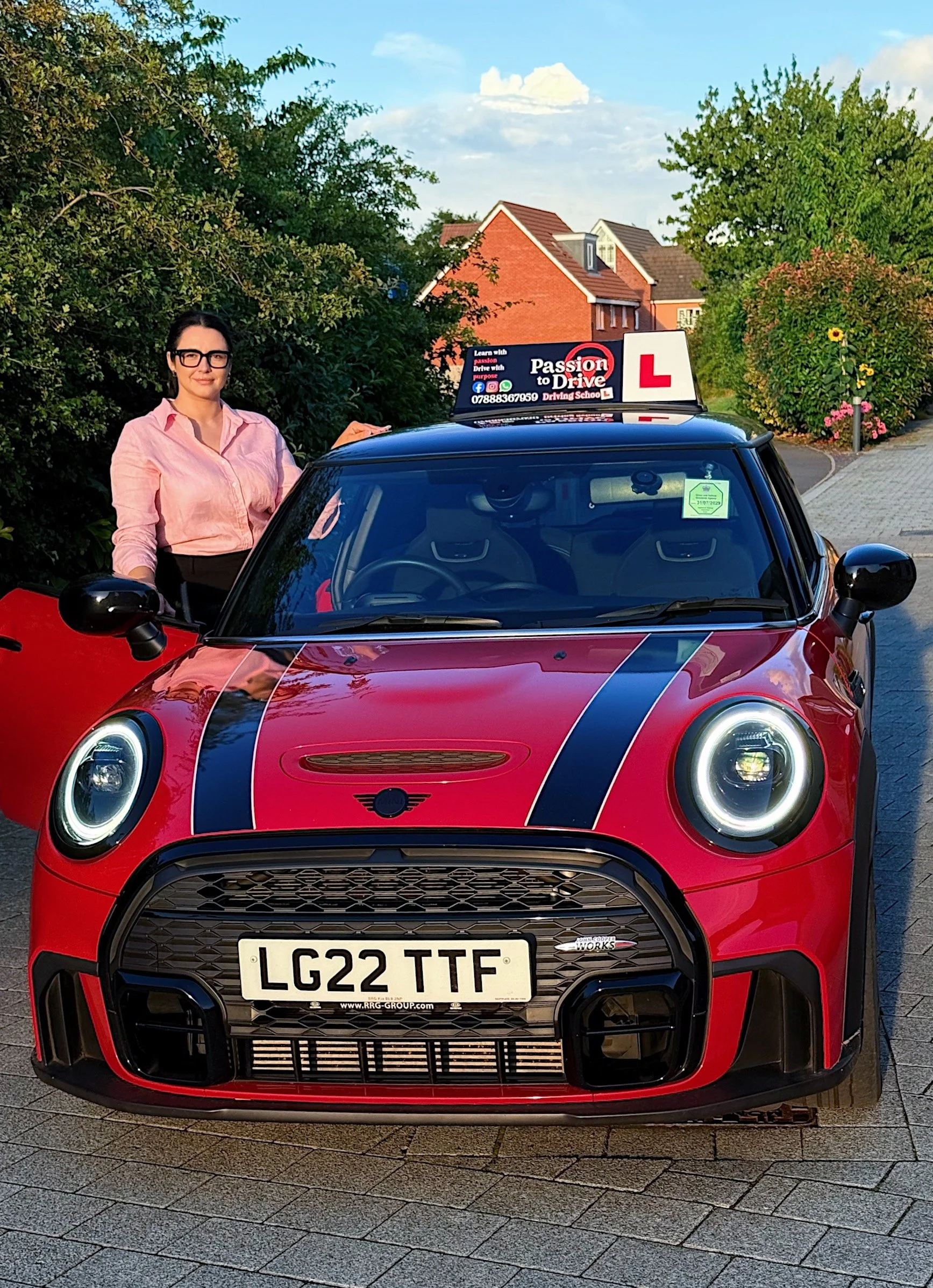 A woman standing next to a red Mini Cooper with a black racing stripe, parked on a residential street. The car has a driving school sign on top, with an 'L' learner symbol, and a sign that says 'Passion to Drive.' The woman is wearing glasses and a pink shirt.