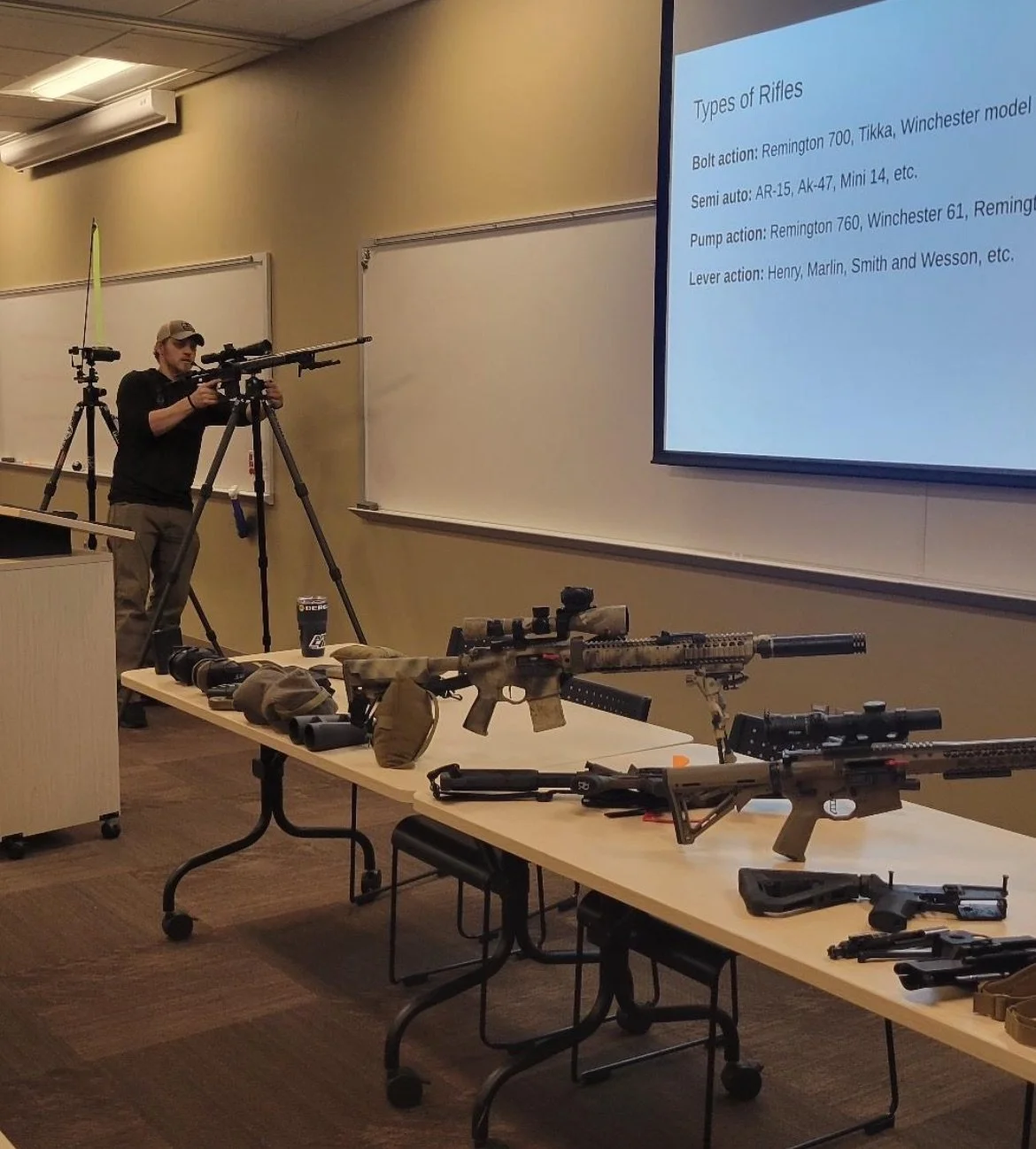 Classroom setting with firearms on tables, a person adjusting a rifle on a tripod, and a presentation slide about types of rifles projected on a screen.