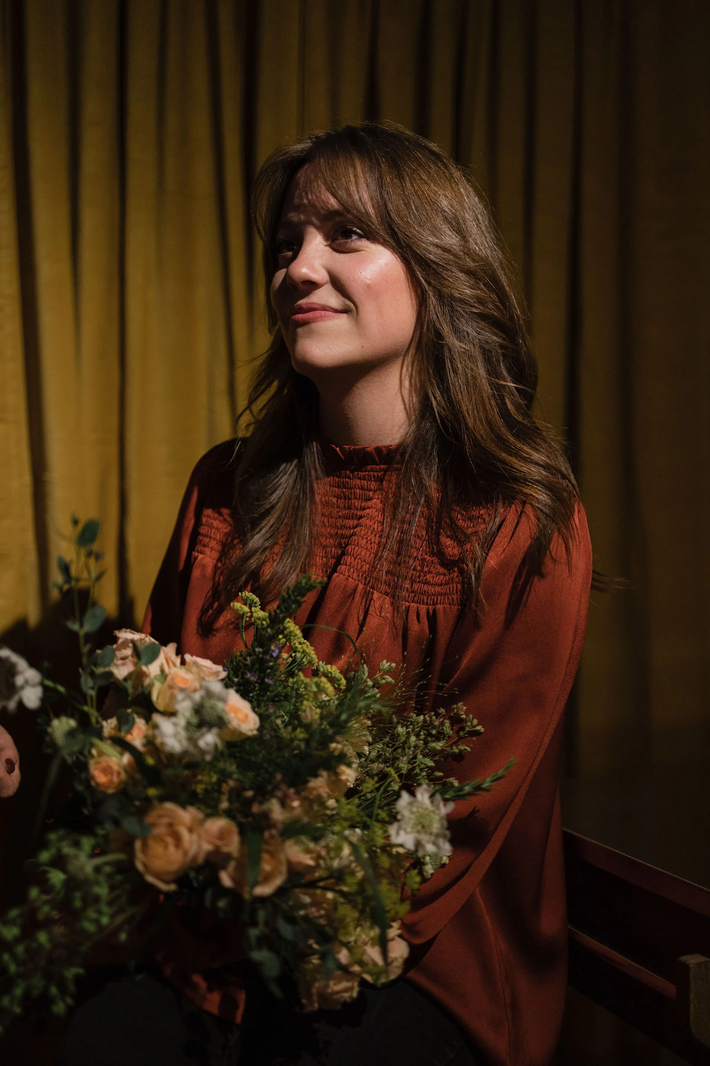 A woman with brown hair sitting in front of a yellow curtain holding a bouquet of flowers.