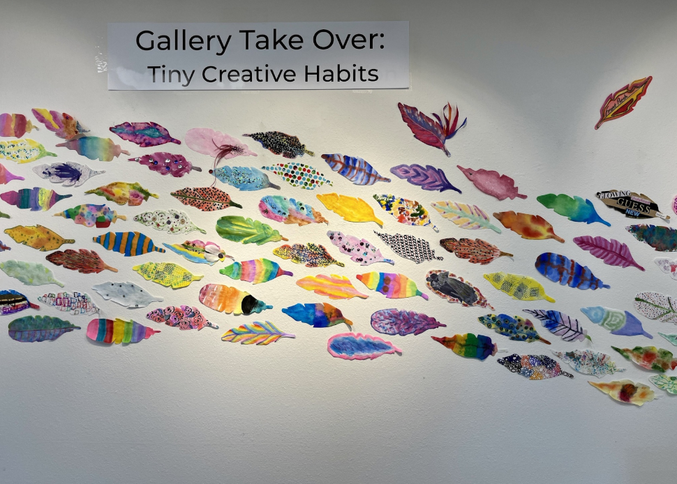 Wall display titled "Gallery Take Over: Tiny Creative Habits" featuring numerous colorful, paper fish art pieces arranged in a pattern.