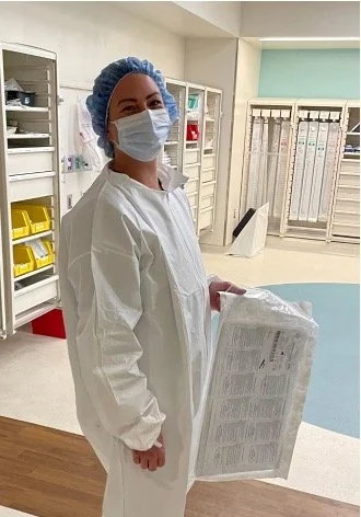 Healthcare worker in scrubs, mask, and hair cover holding a medical tray in a hospital setting.