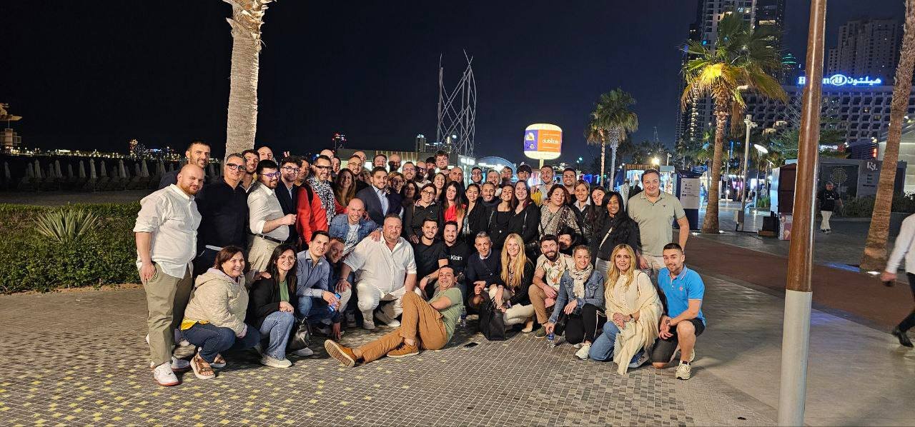 gruppo di persone in piedi e sedute in una zona pedonale di sera, con luci e palme sullo sfondo, in un'area urbana