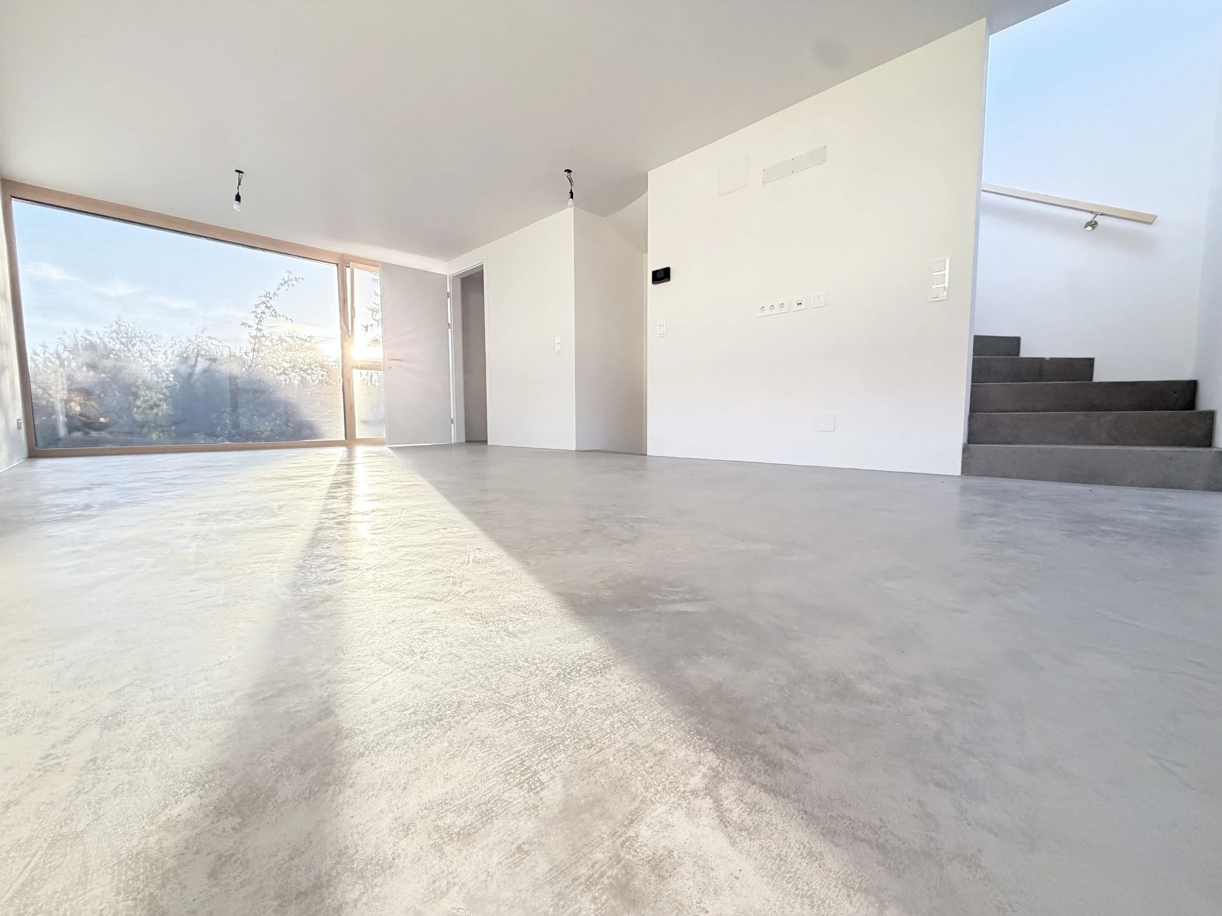 Leeres, helles Wohnzimmer mit großen Fenstern und moderner Treppe.