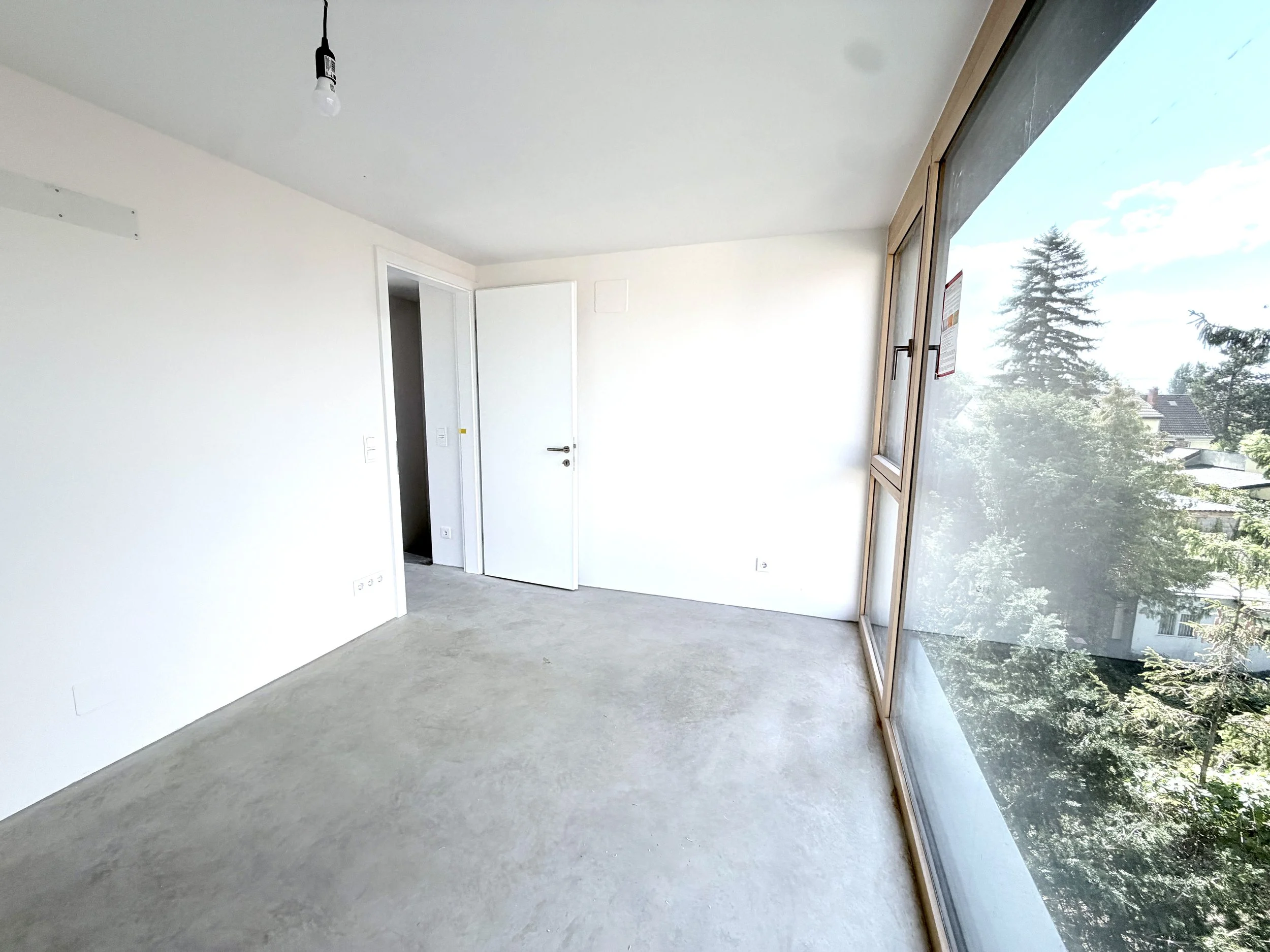 Ein leeres, helles Zimmer mit weißen Wänden, einem offenen Tür und einem großen Fenster, das auf Bäume und den Himmel hinausgeht.