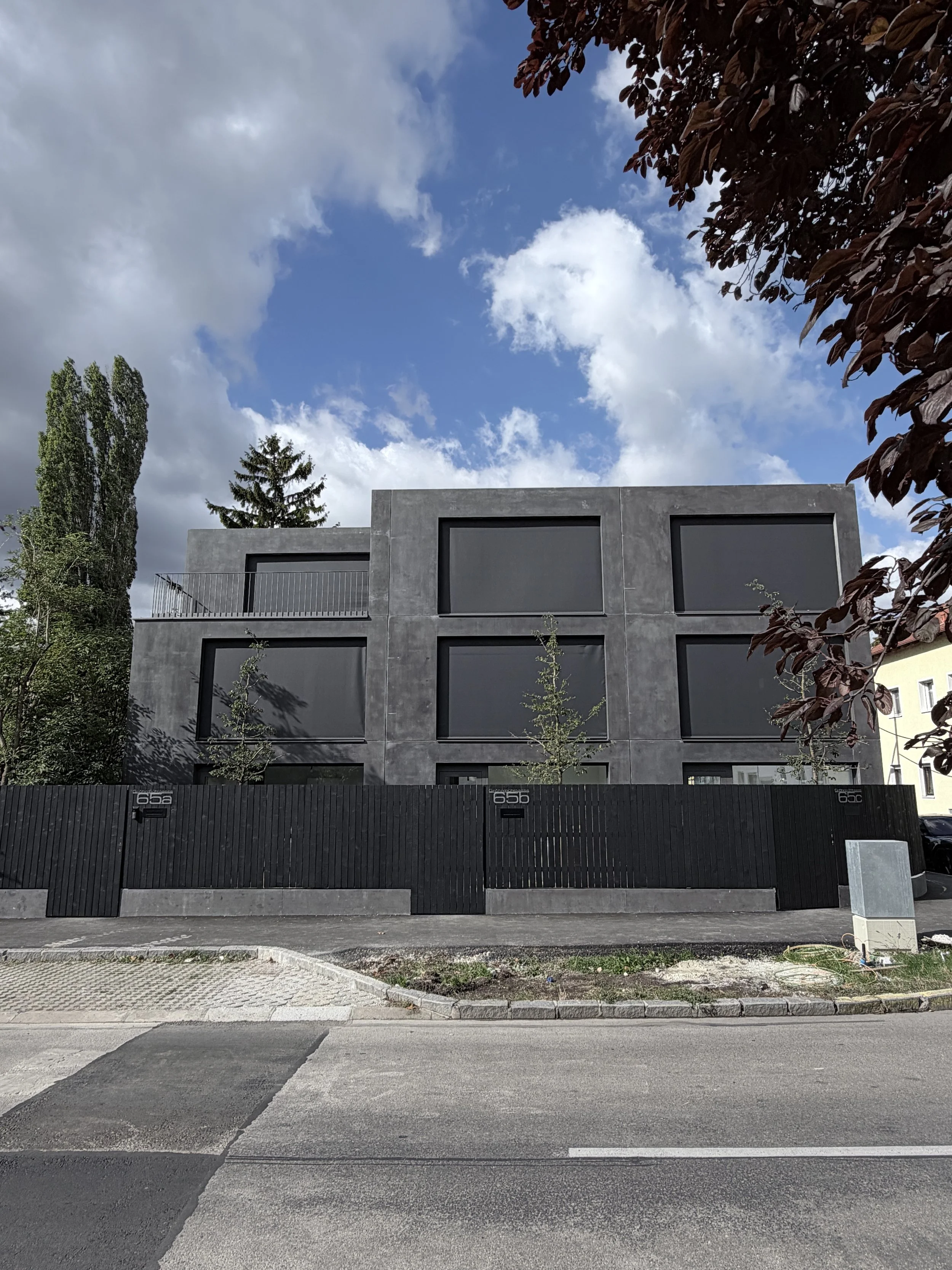 Moderner grauer Gebäudekomplex mit großen, schwarzen Fenstern und einem schwarzen Zaun davor. Der Himmel ist teils bewölkt, umgeben von Bäumen.