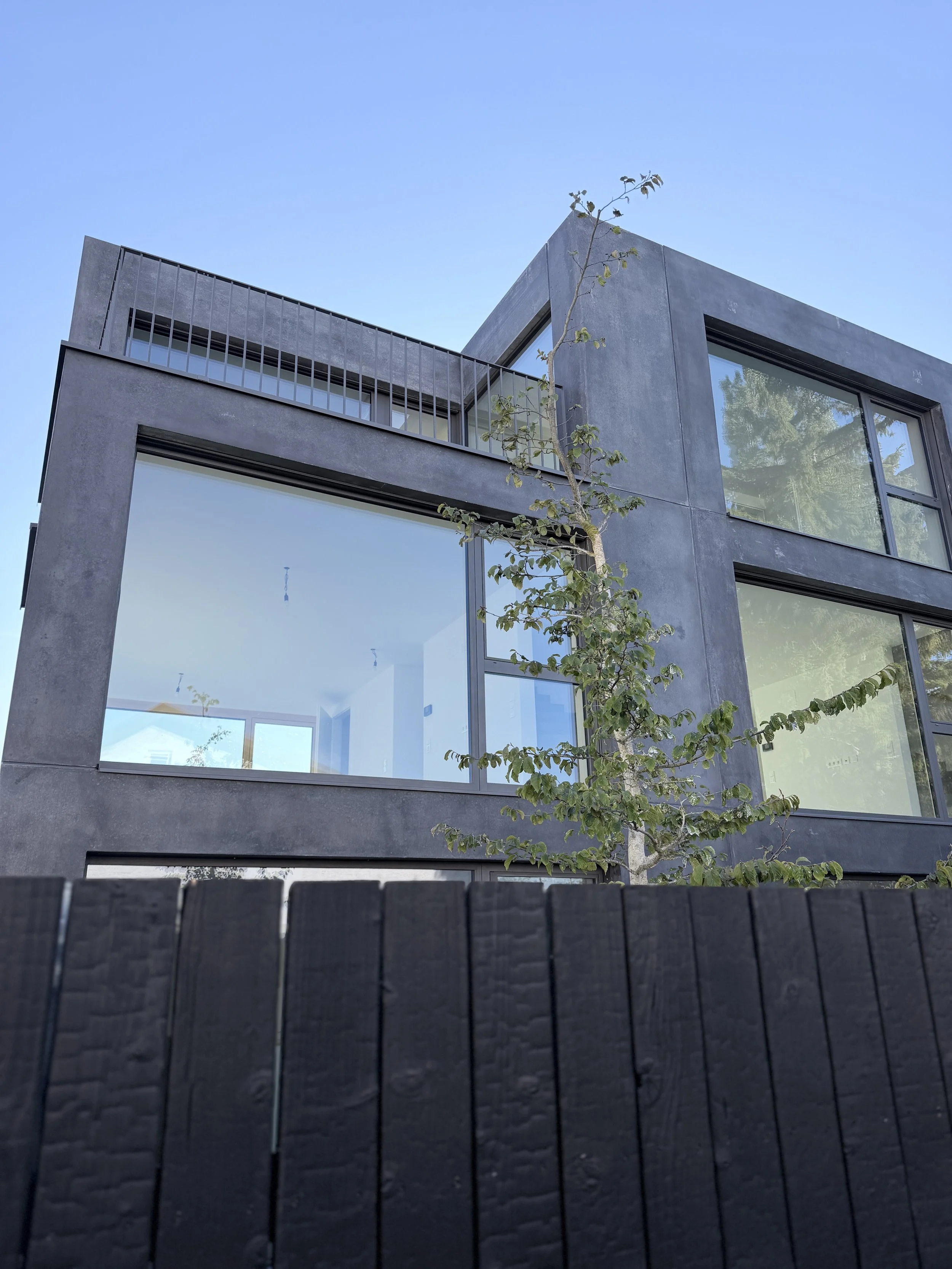 Moderne graue Hausfassade mit großen Fenstern und einem kleinen Baum vor dem Haus, das im Himmel reflektiert.