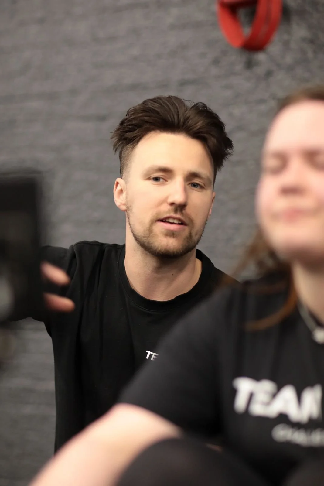 A young man with brown hair and a beard is smiling slightly, wearing a black shirt, and appears to be taking a selfie or is posed for a photo. There is a woman with brown hair and a black shirt in front of him, but she is out of focus.