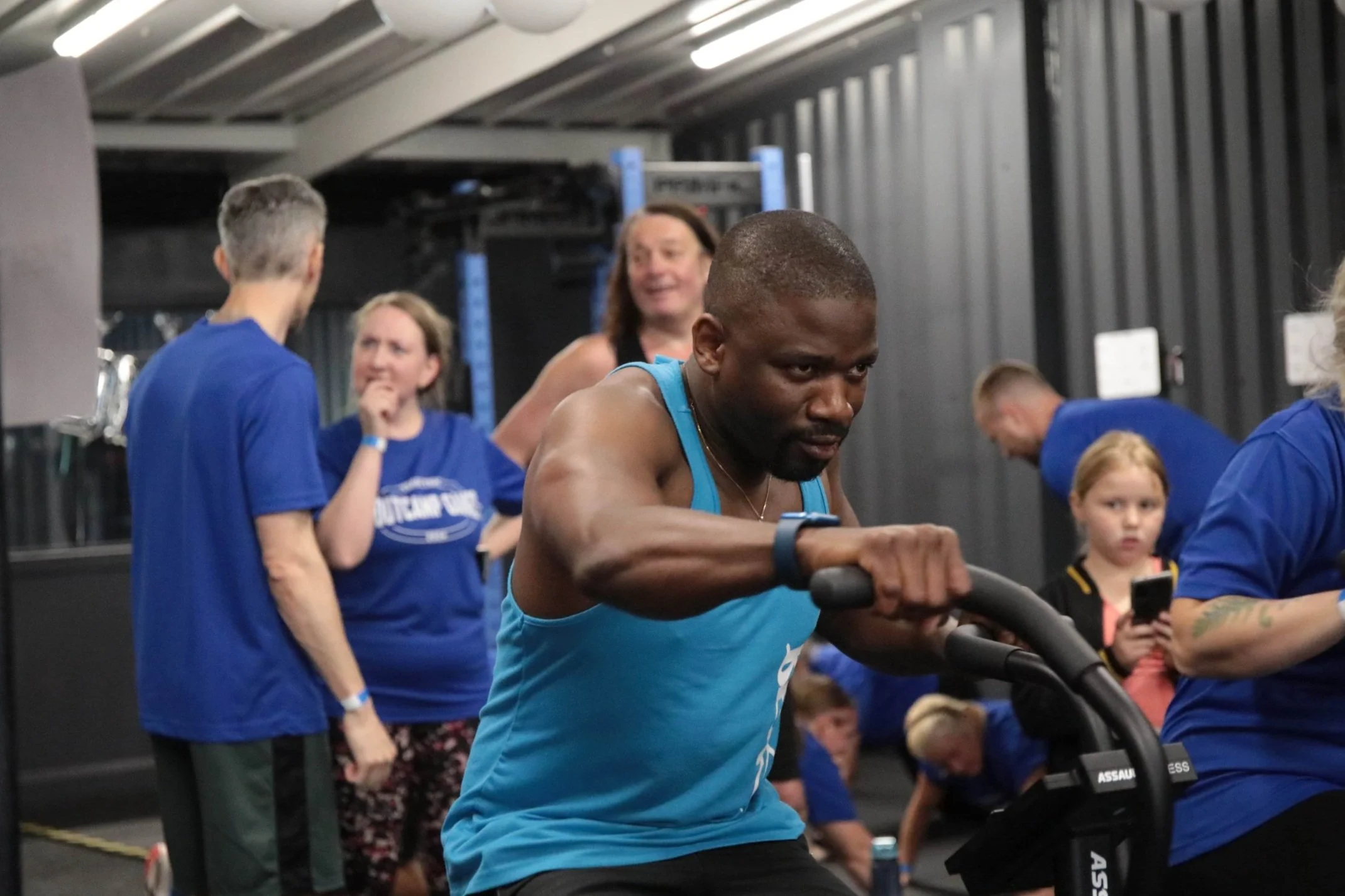 A man in a blue tank top works out on a rowing machine at a gym while several other people, including women and children, talk and observe in the background.