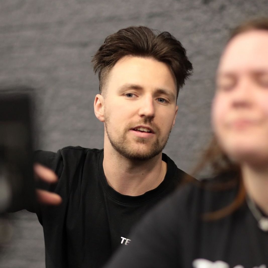 Young man with dark hair and beard taking a selfie with a blurred woman in the foreground. The background appears to be a gray wall.