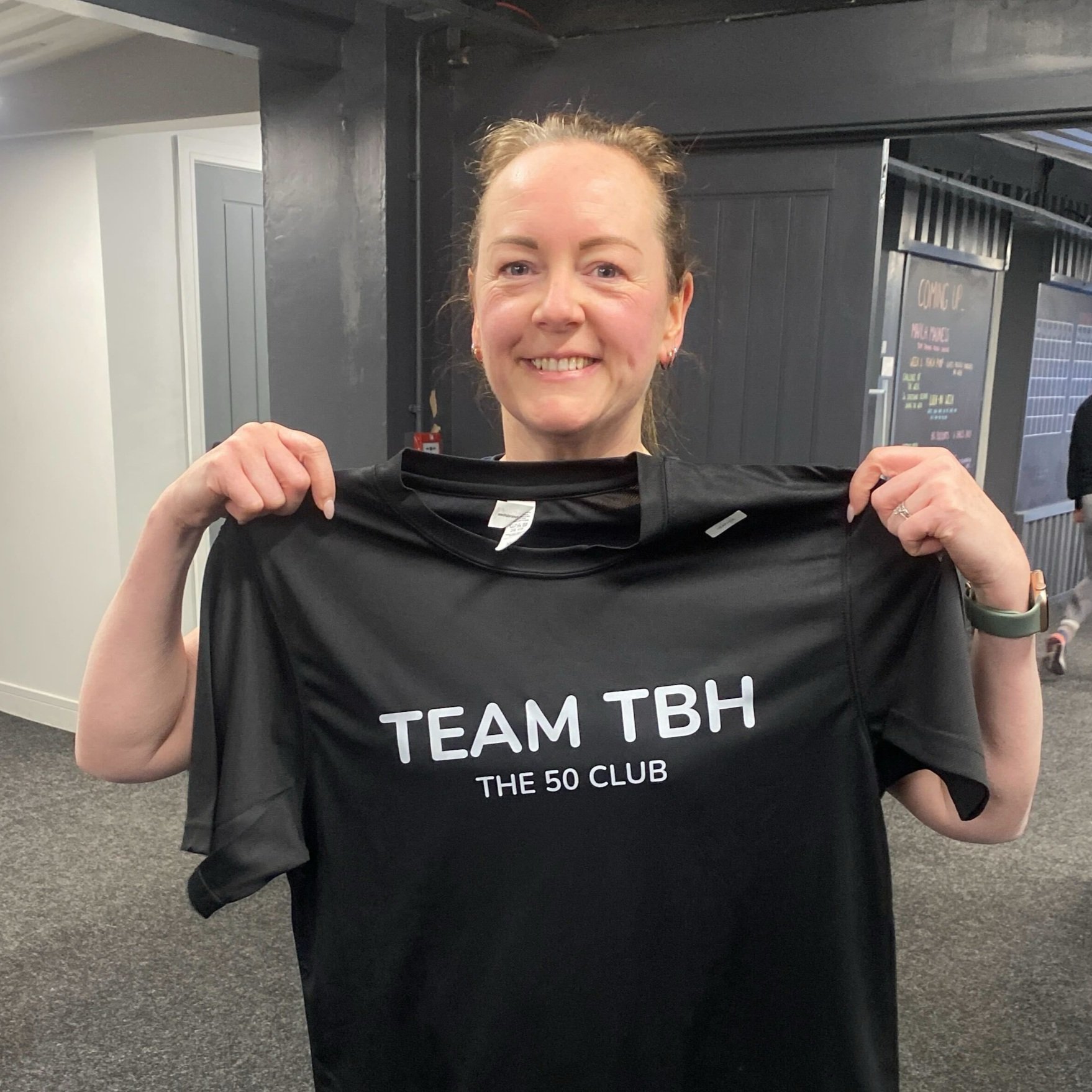 A woman smiling and holding a black T-shirt that says 'TEAM TBH THE 50 CLUB' in white letters.
