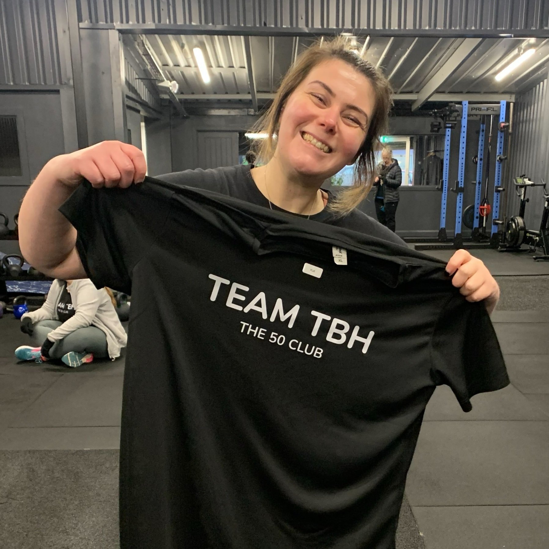 A woman in a gym holding a black T-shirt that says "TEAM TBH THE 50 CLUB" and smiling at the camera.