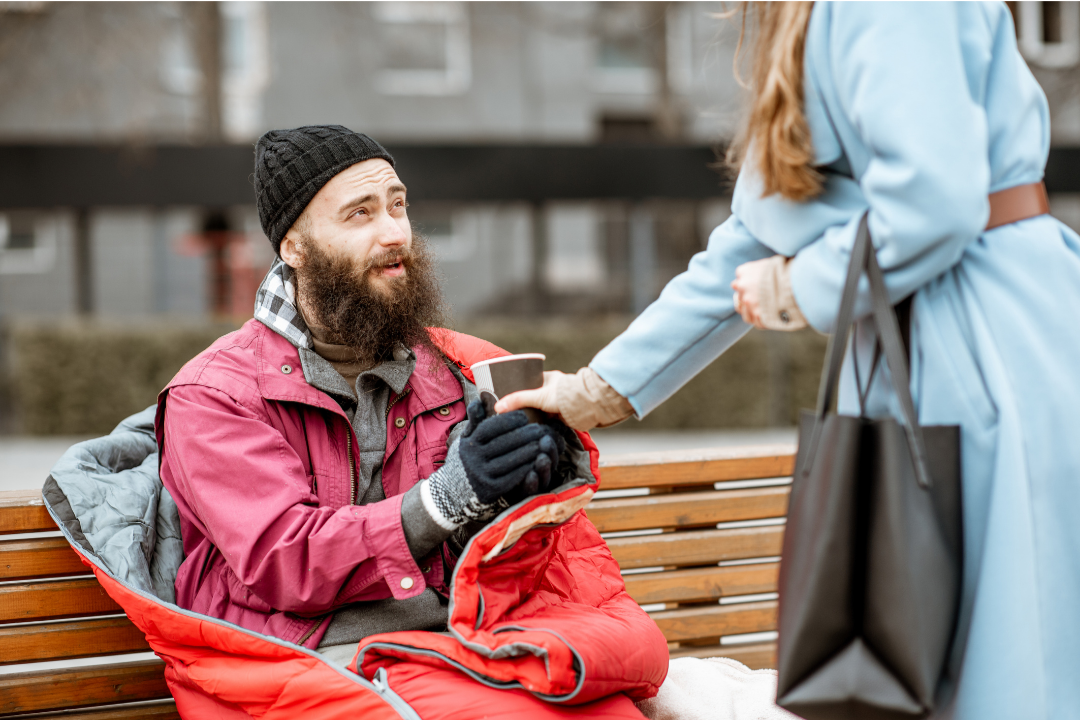 A homeless man with a beard and wearing a black beanie, red jacket, and gloves sits on a park bench. A woman in a light blue coat offers him a cup.