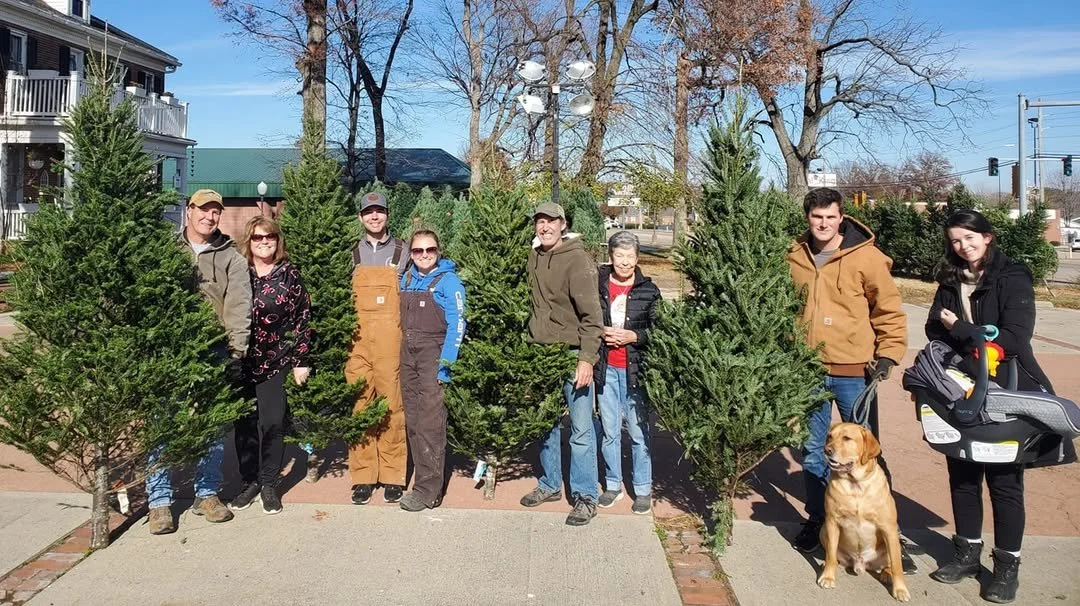 A group of nine people and a dog standing outdoors behind a row of Christmas trees at the Waterloo Optimist Club Christmas Tree Lot, enjoying a sunny day with leafless trees and a sidewalk in the background.