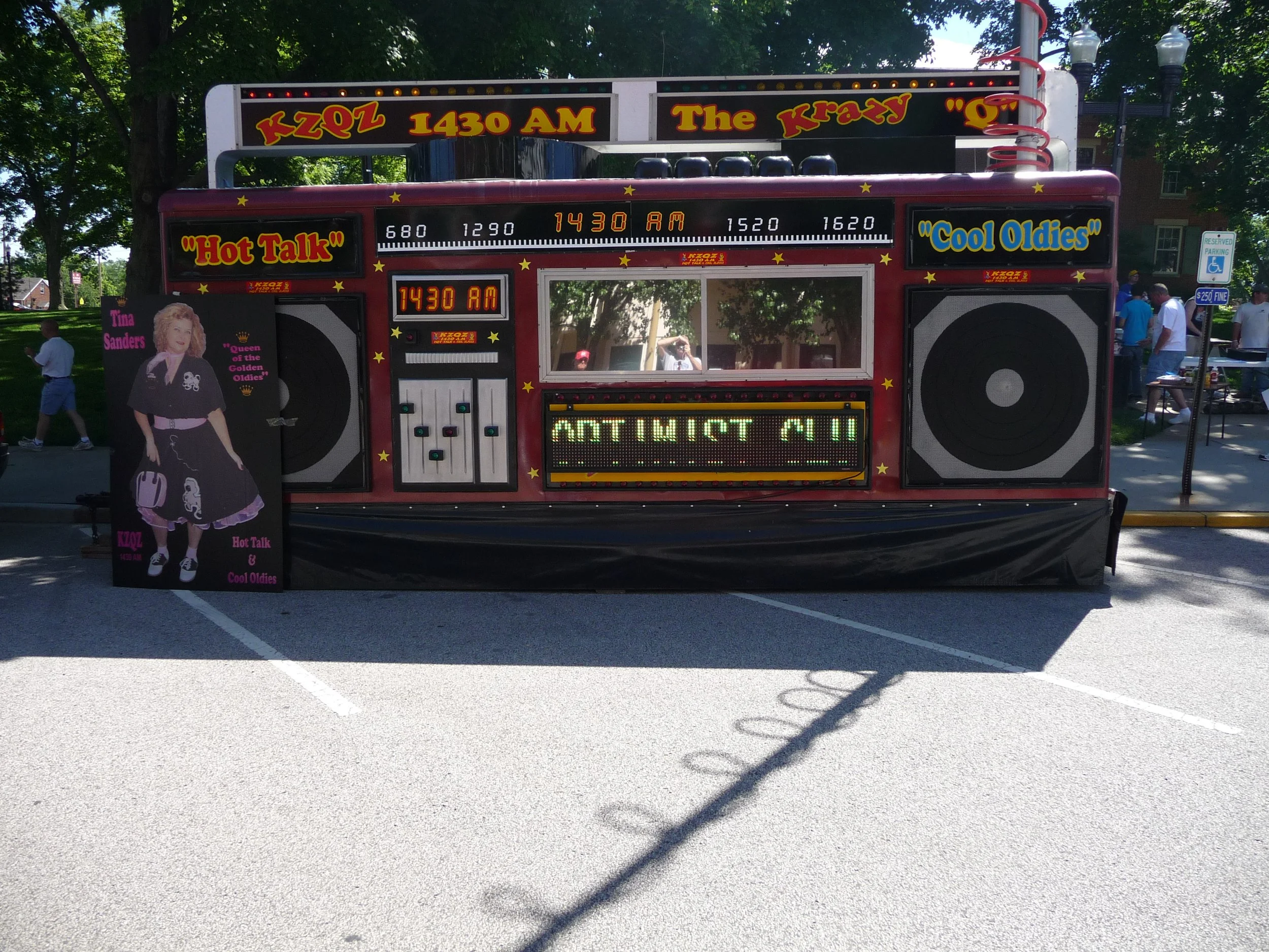 A vintage-style radio-themed promotional booth for a radio station, decorated with colorful signs and a large speaker, displaying show times and slogans at the Waterloo Optimist Club car show, with a standee of a woman in a black dress and pink text.