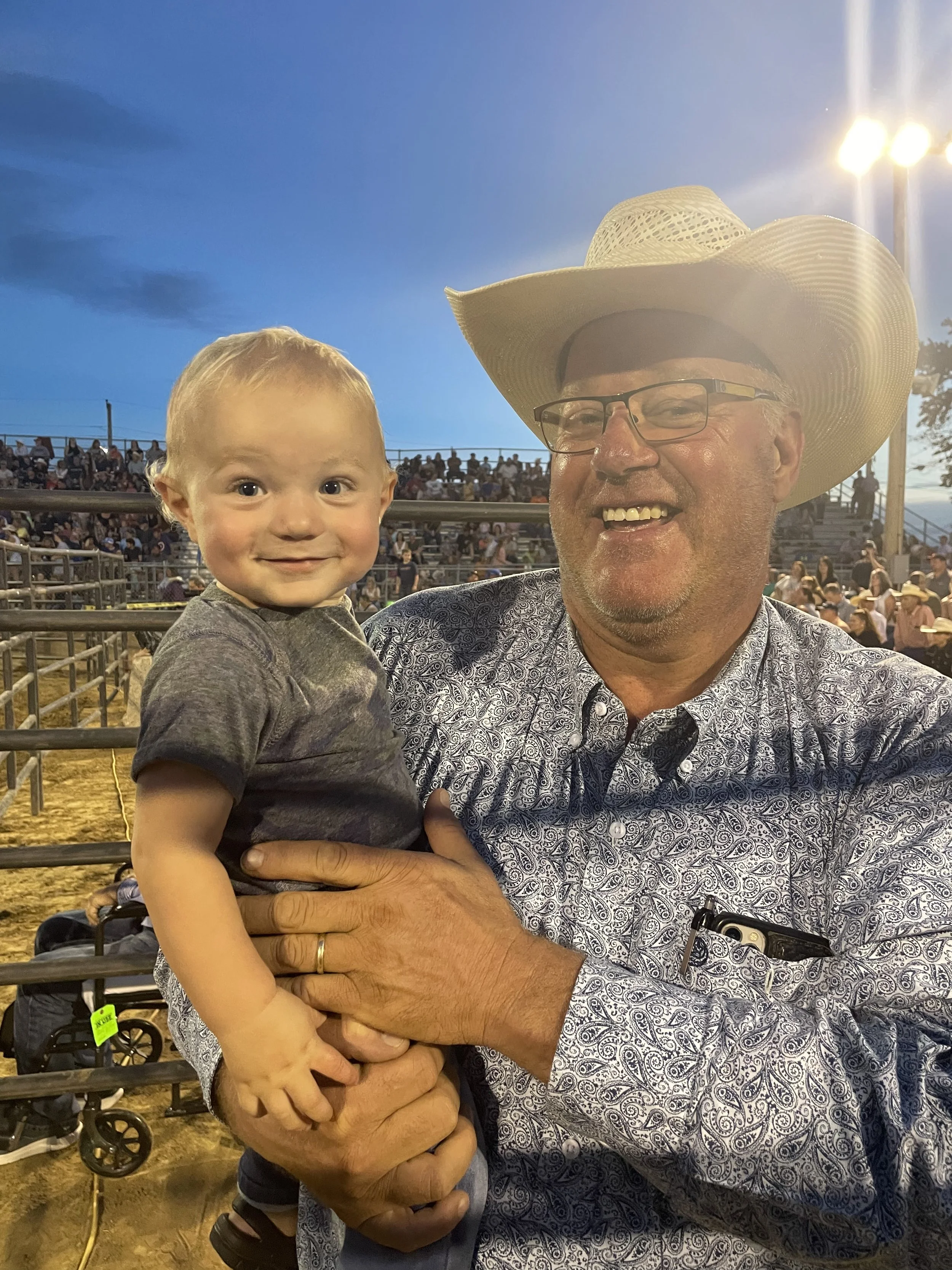 A man wearing a cowboy hat and glasses holding a young boy in a gray shirt at the Blue Army Bull Bash during dusk. The background shows a crowd sitting on bleachers and bright stadium lights illuminating the scene.
