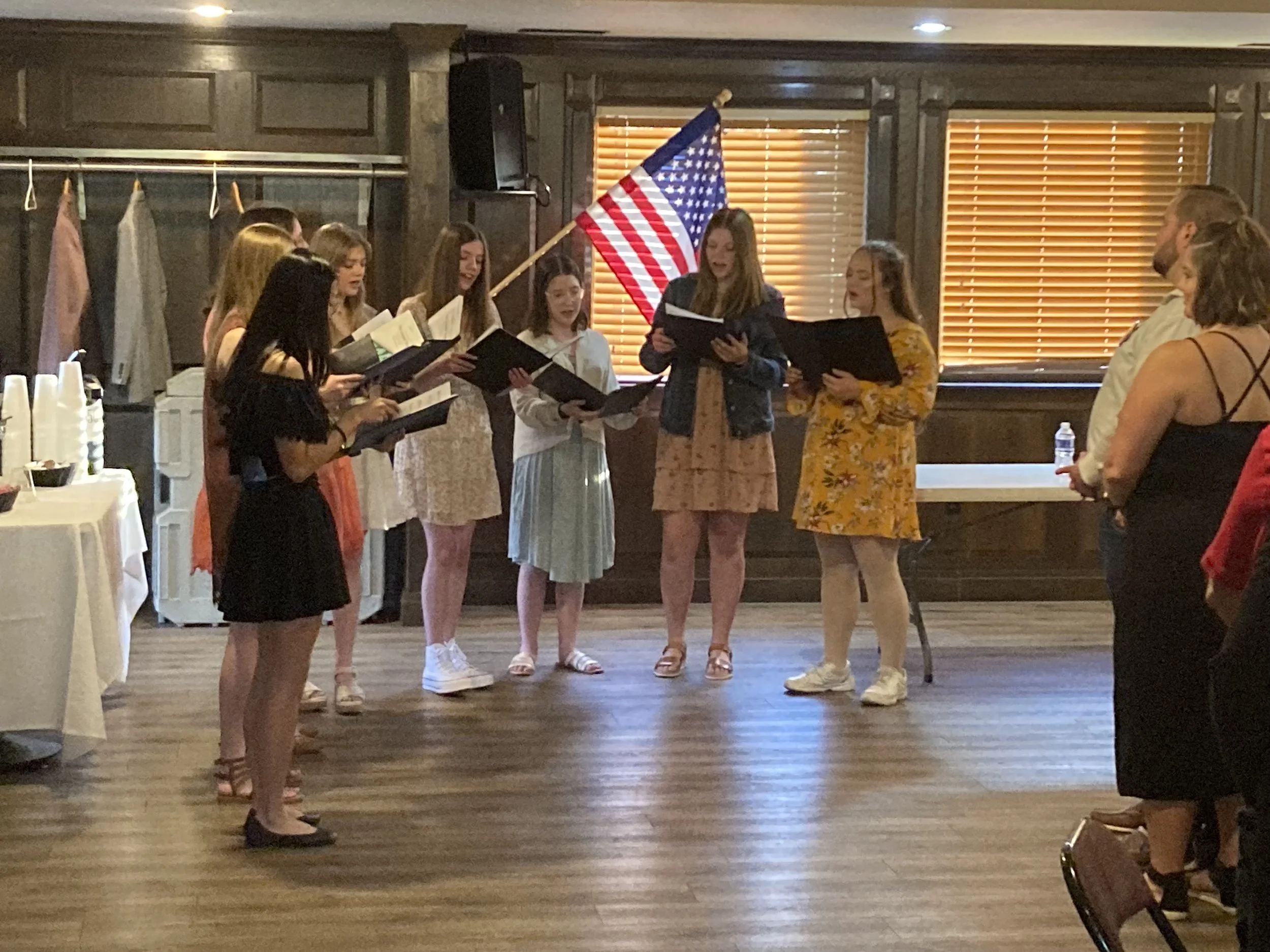 Group of young women singing in a room with wooden walls, an American flag, and window blinds, while a man and woman listen.