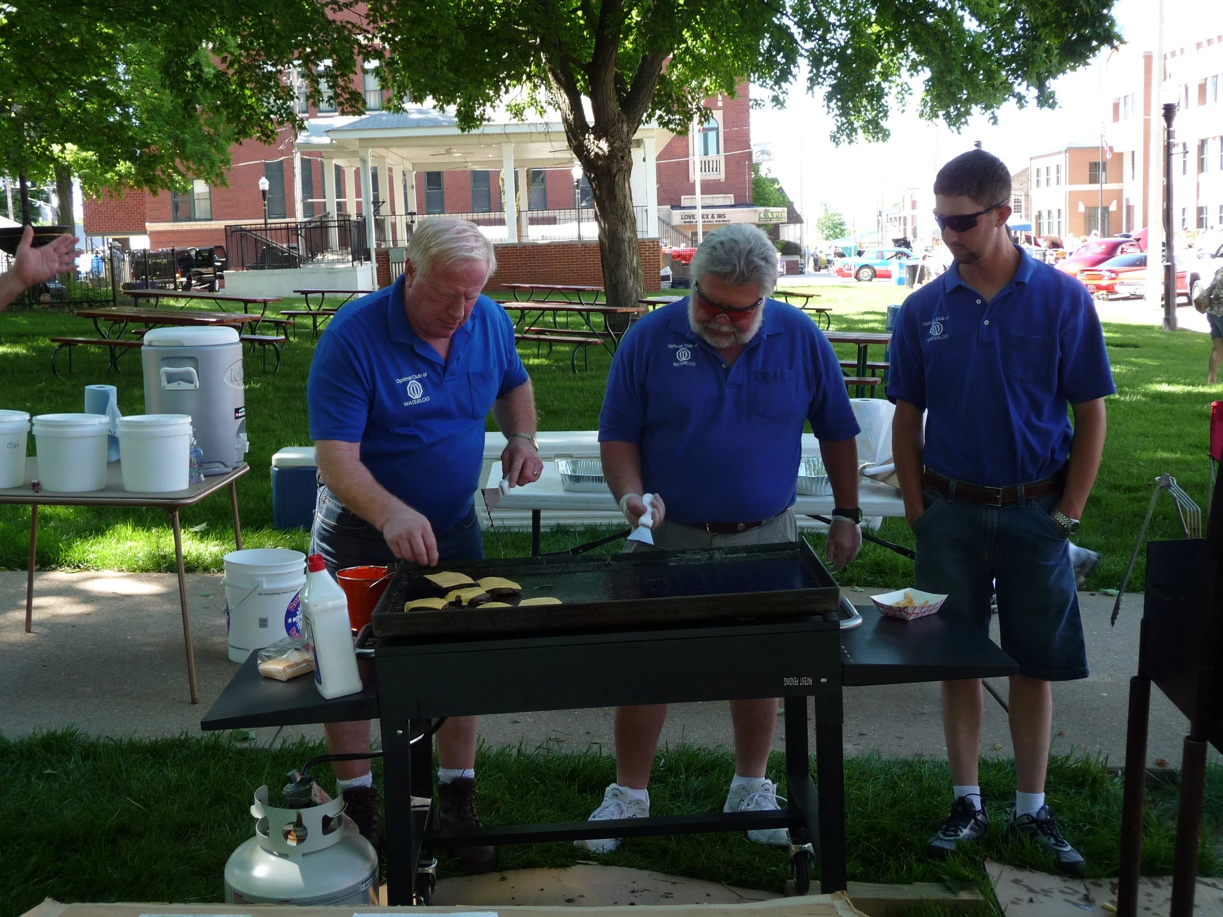 Three Waterloo Optimist Club members in blue shirts cooking burgers on an outdoor grill at a park, with tables and a building in the background.