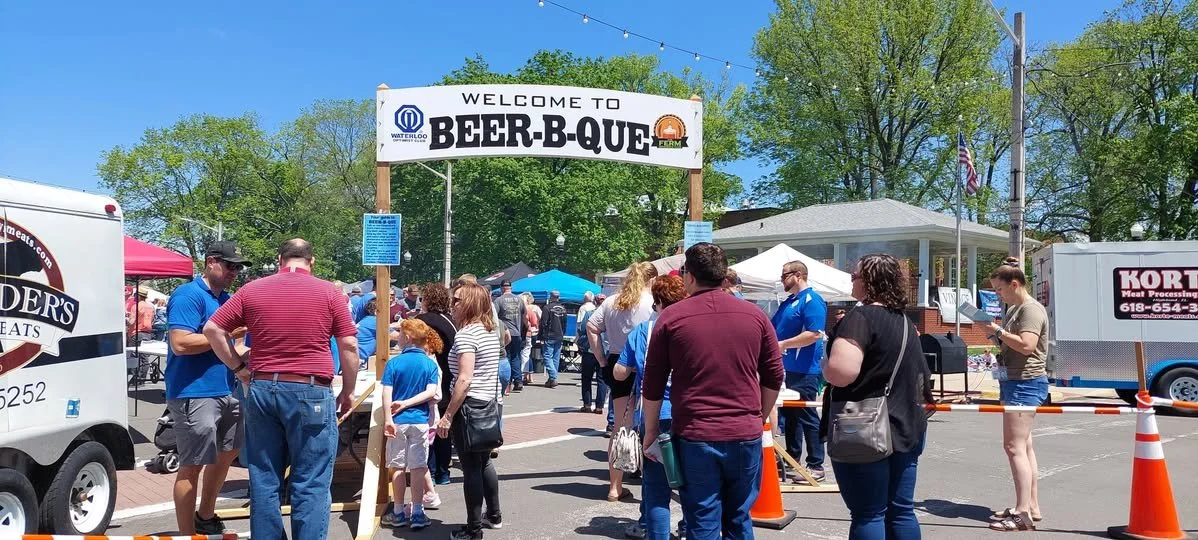 People waiting in line at a beer barbecue event with tents and food trucks, under a clear blue sky, with a sign welcoming to Beer B Que.