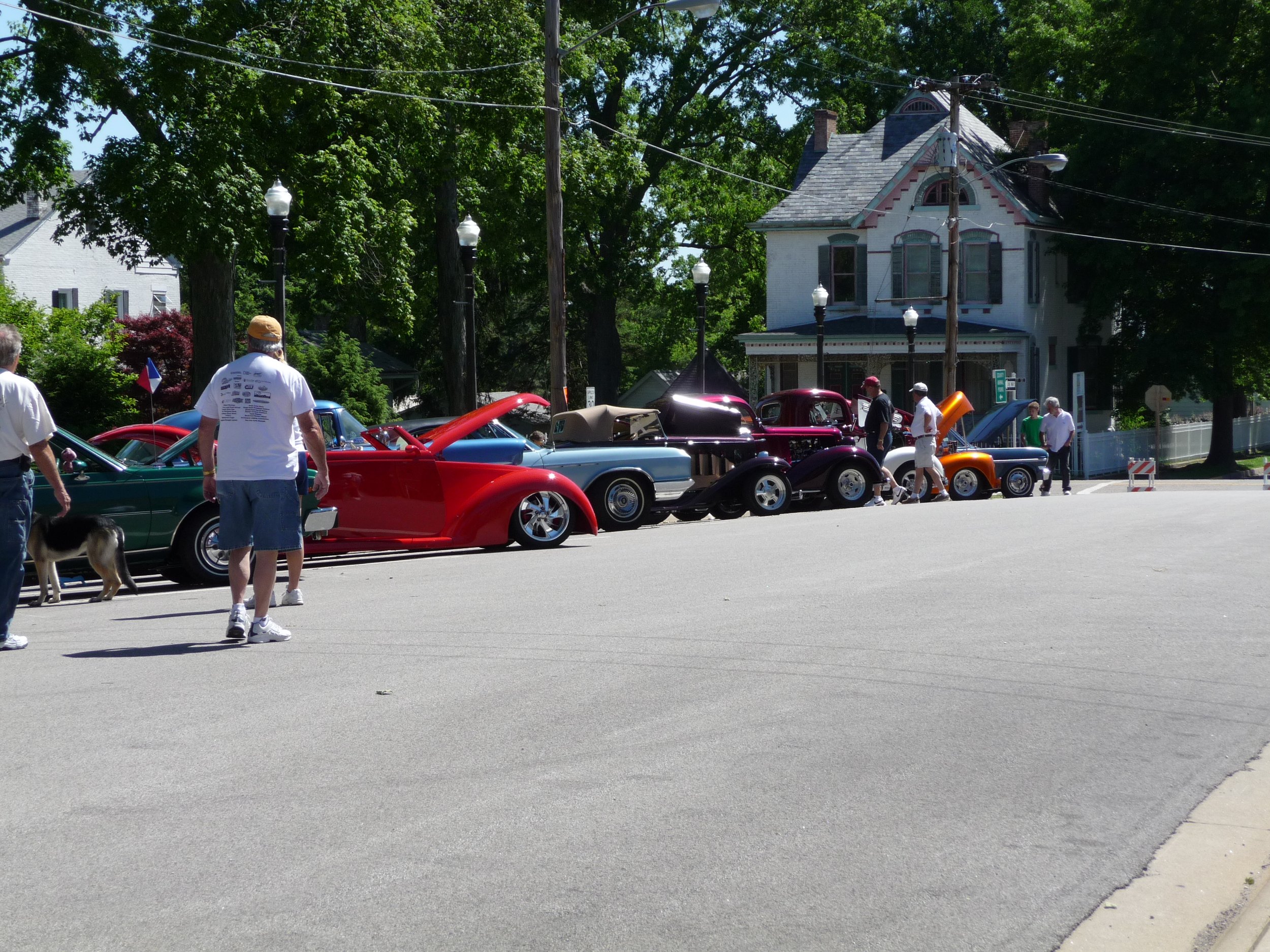 A row of classic cars on display during the Waterloo Optimist Club car show, with people walking and looking at the vehicles, in front of a residential house with trees and street lamps.