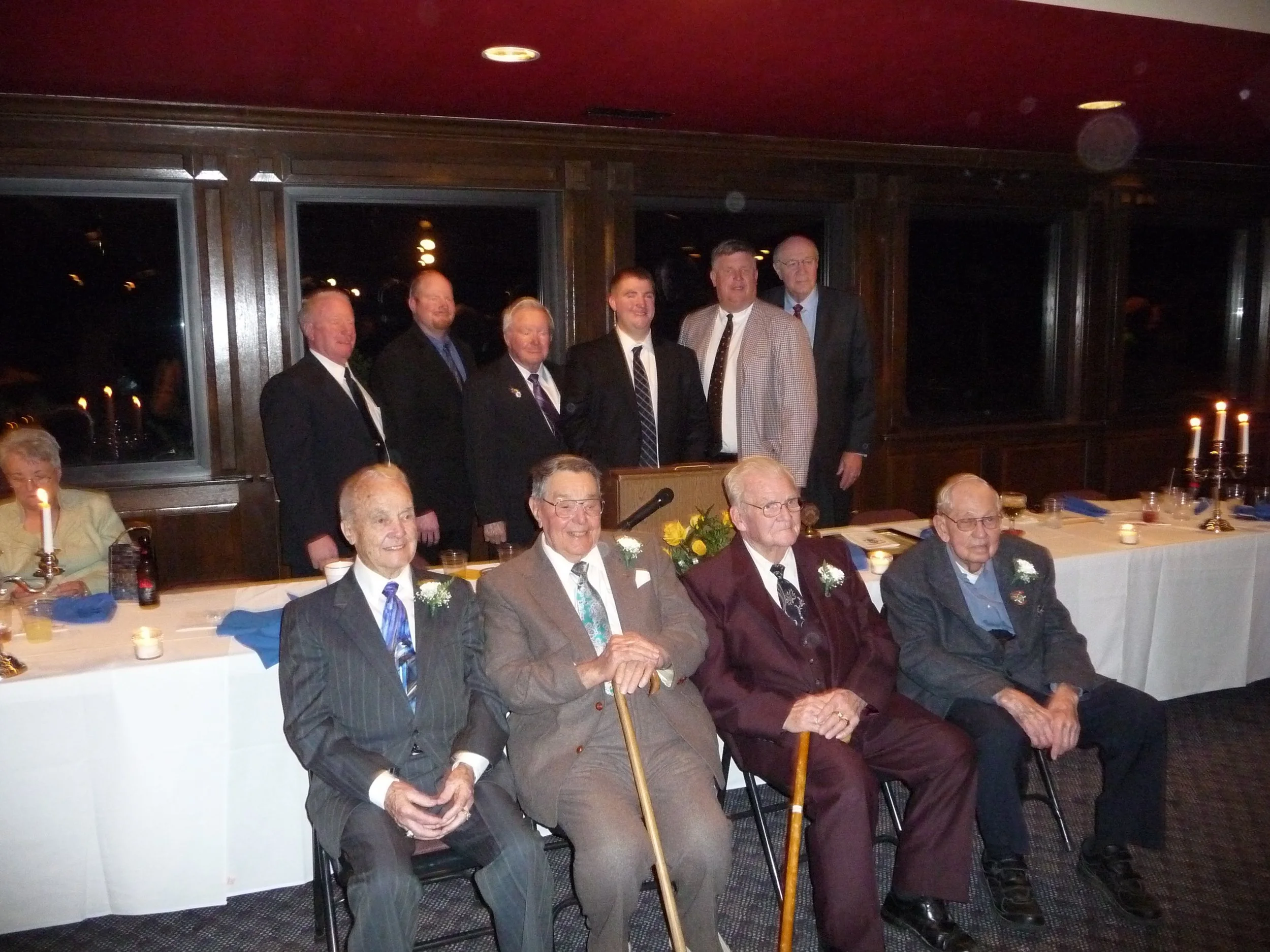 A group of elderly men and women in formal attire gathered at a dinner event, with some seated at a table and others standing behind them in a dimly lit room with dark wood paneling and candles.