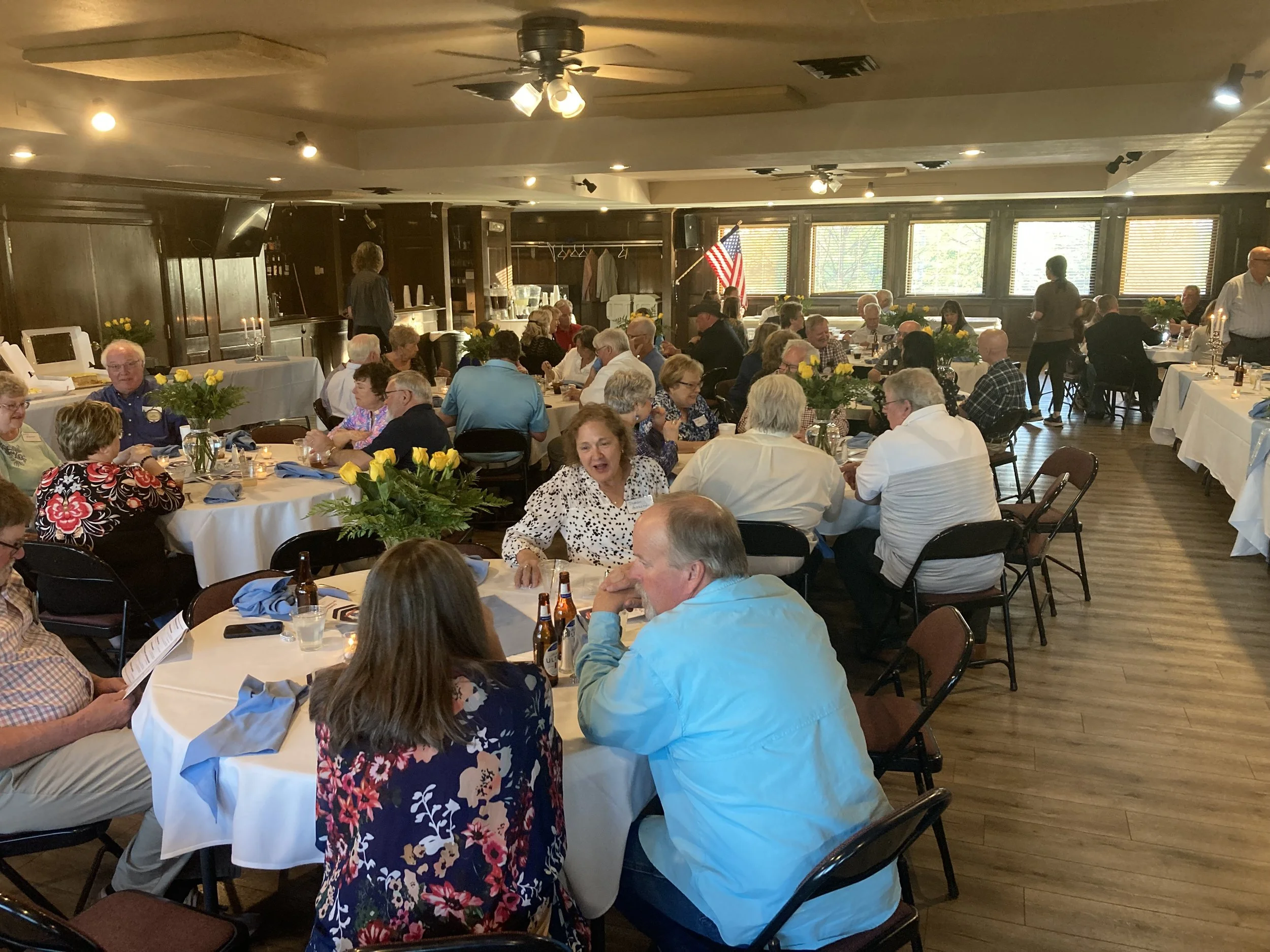 Guests seated around tables in a banquet hall during an event, with floral centerpieces, some tables have candles, and a stage area in the background with an American flag.