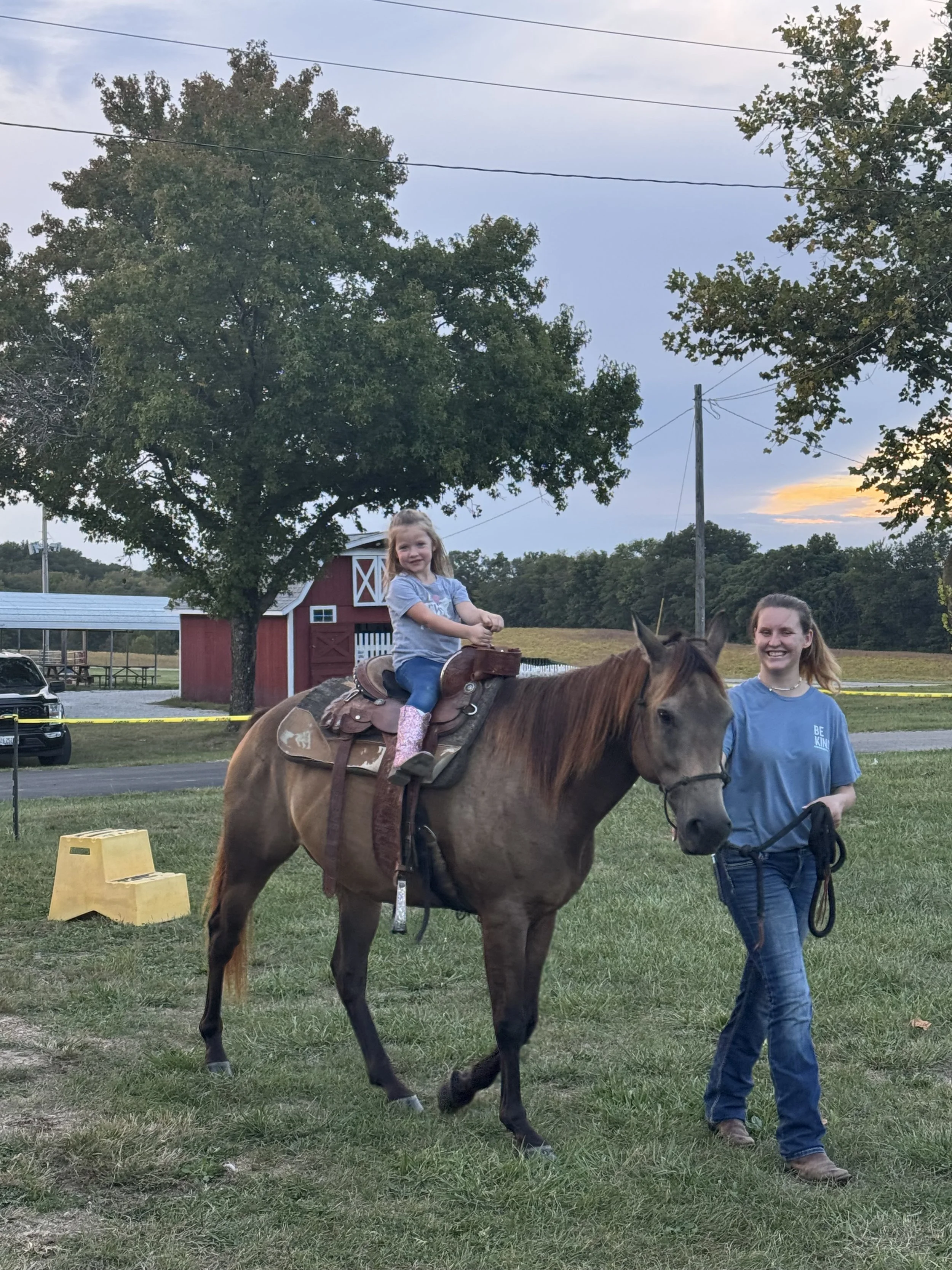 A girl riding a horse with a woman walking beside her in a grassy outdoor area at sunset, with trees and a red barn in the background at the Blue Army Bull Bash.