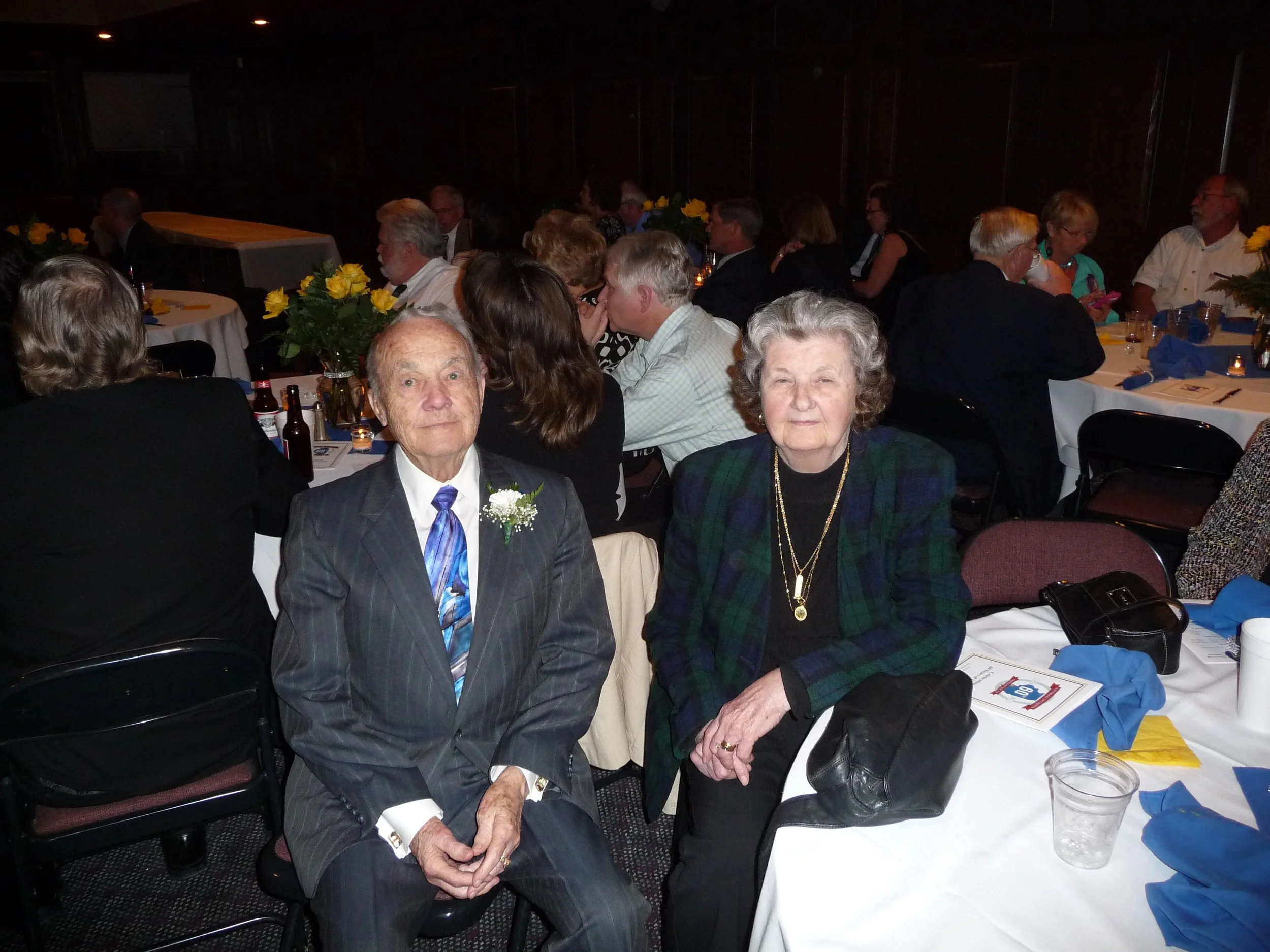 An elderly man and woman seated at a dinner table during a formal event, with other guests in the background and floral centerpieces on the tables.