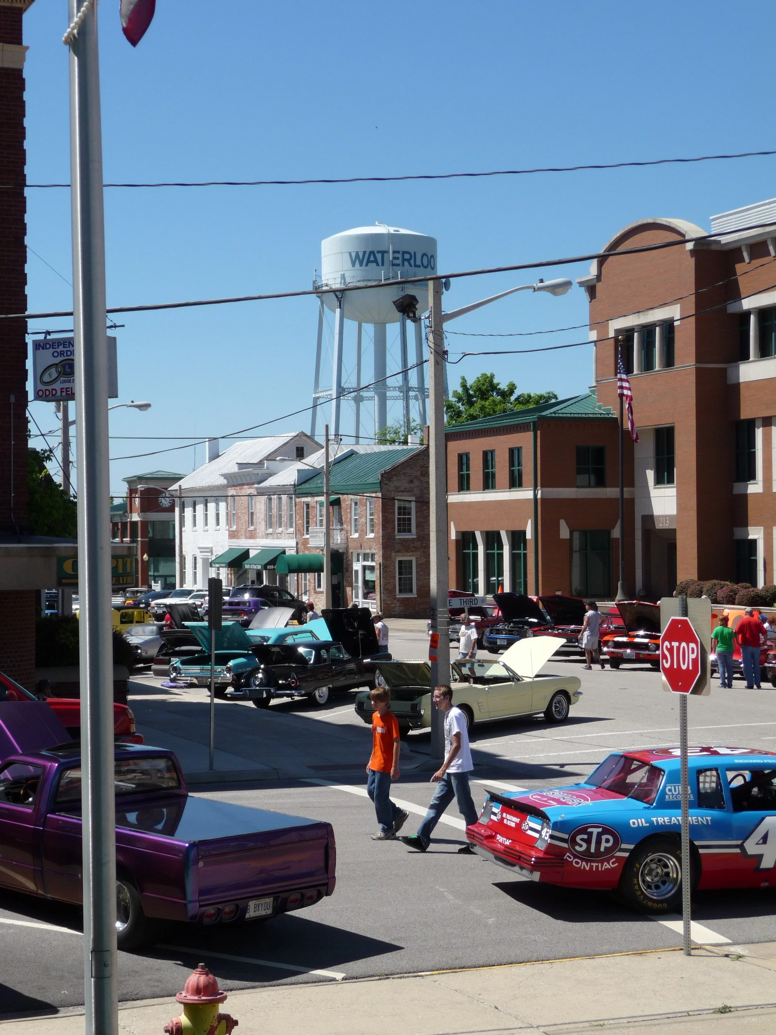 A street scene showing the Waterloo Optimist Club car show with vintage cars parked along the street, people walking, and a water tower that reads 'WATERLOO' in the background. There are buildings, a stop sign, and a hydrant visible.