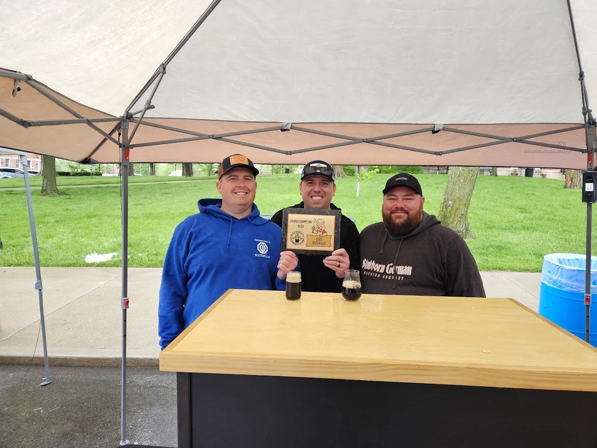 Three men standing behind a wooden counter under a canopy, smiling at the camera at the Waterloo Optimist Club Beer-B-Que There are two drinks in front of them on the counter. A grassy park with trees is in the background.