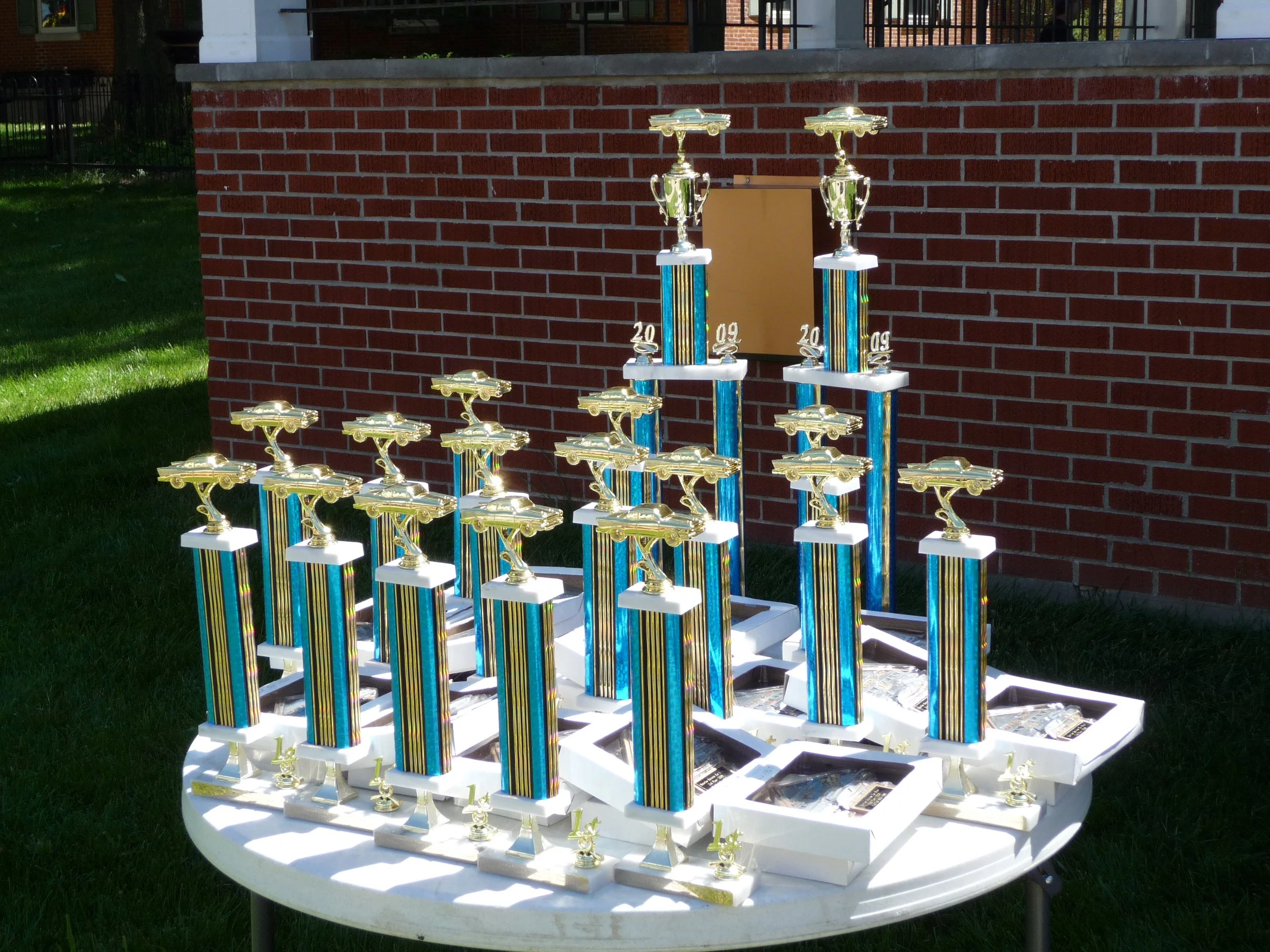 Multiple Waterloo Optimist Club car show trophies with gold-colored race cars on top, arranged on a white round table outdoors against a red brick wall.