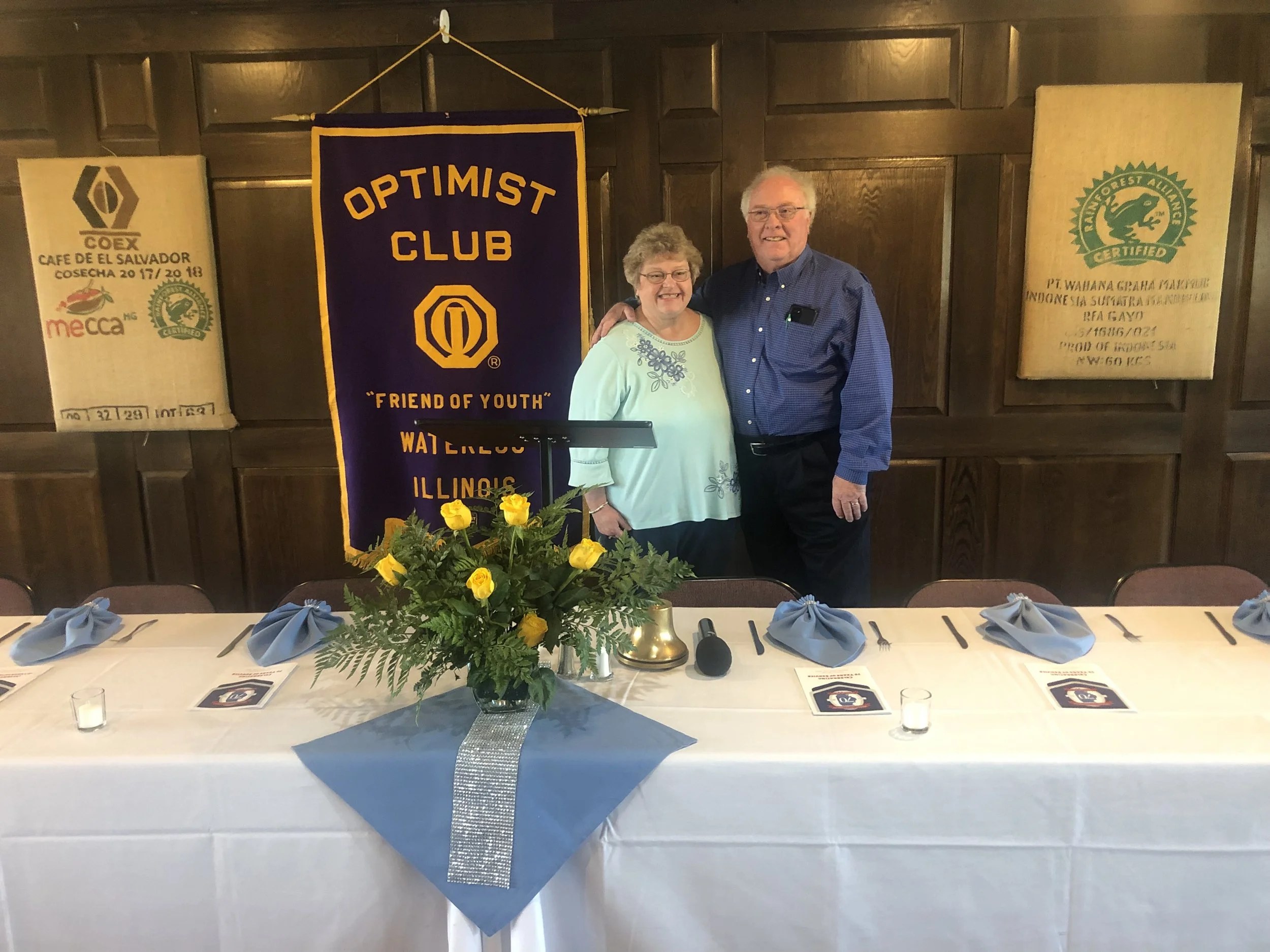 A man and woman standing together at a banquet table, smiling, with a purple Optimist Club banner behind them. The table is decorated with a bouquet of yellow flowers, blue napkins, and dining utensils.