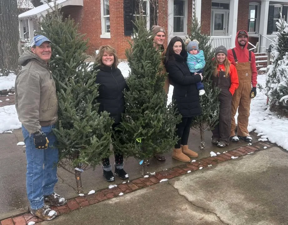 Group of seven people standing outdoors in winter at the Waterloo Optimist Club Christmas Tree Lot, holding Christmas trees, with snow on the ground and a brick house in the background.
