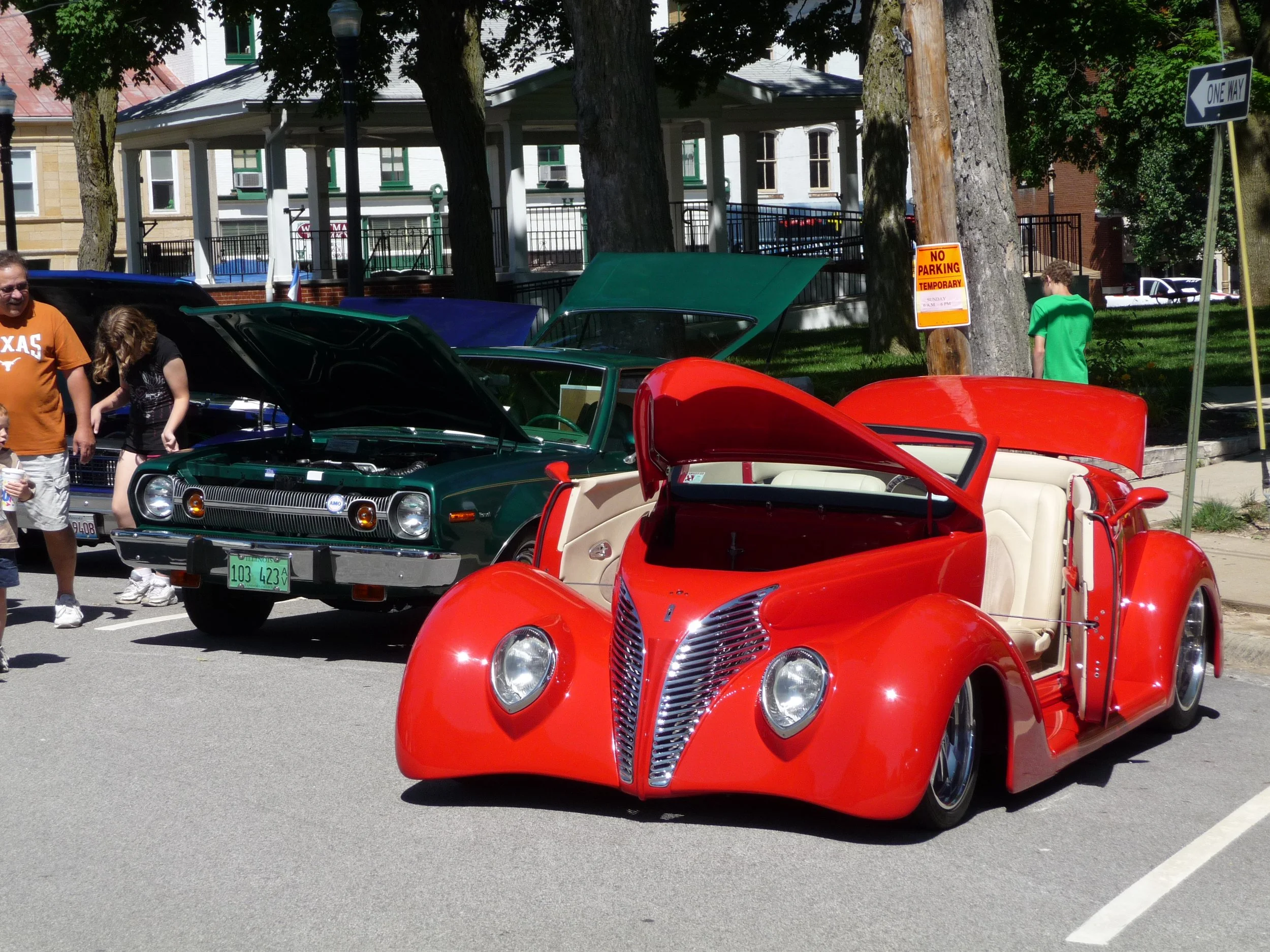 Red vintage convertible car with its hood open on display at the Waterloo Optimist Club car show, with a green vintage car behind it also with its hood open, and several people walking and observing the cars.