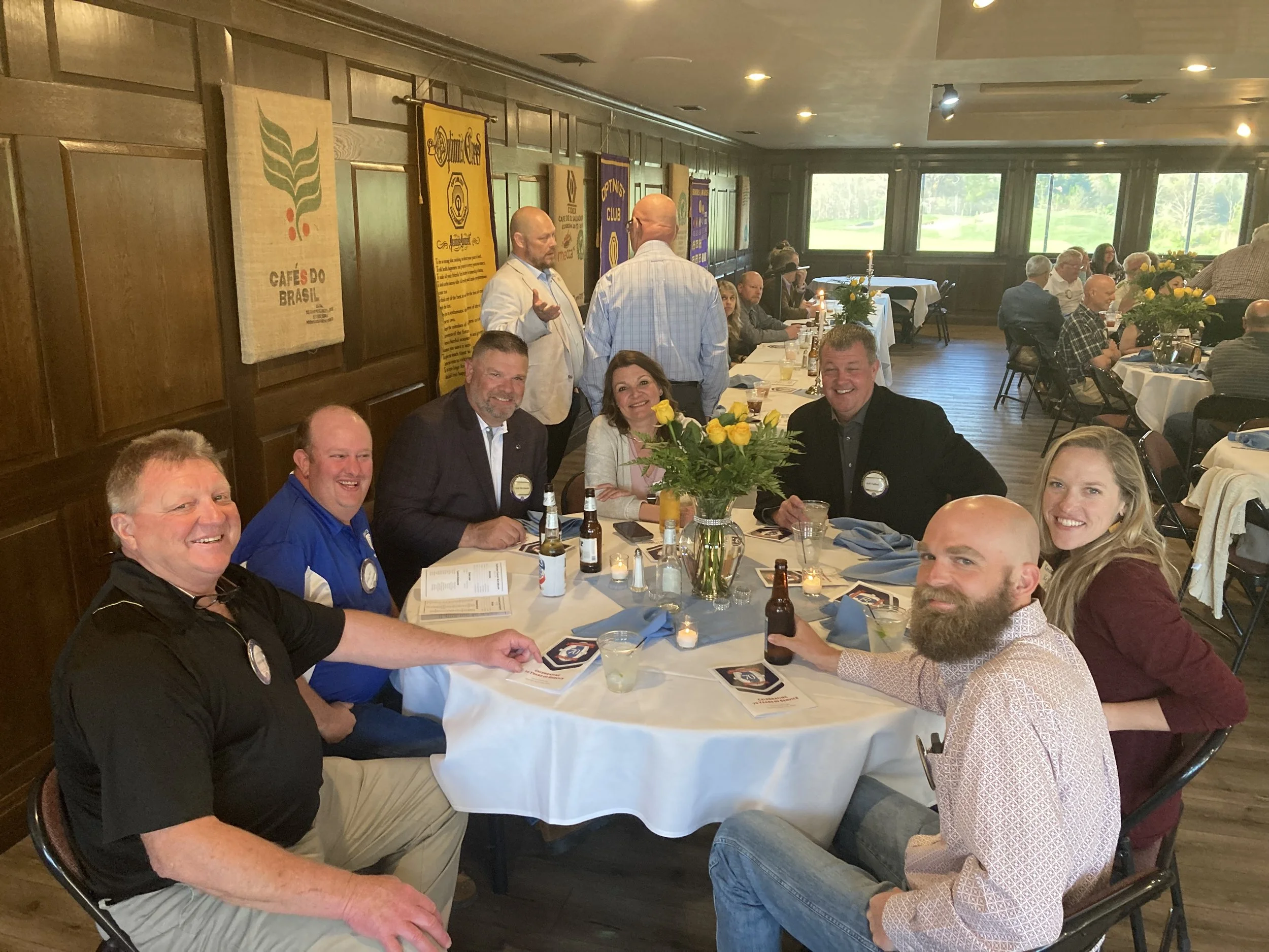 People sitting around a round table at a formal event in a restaurant or banquet hall, with a small flower arrangement in the center, smiling and enjoying drinks.