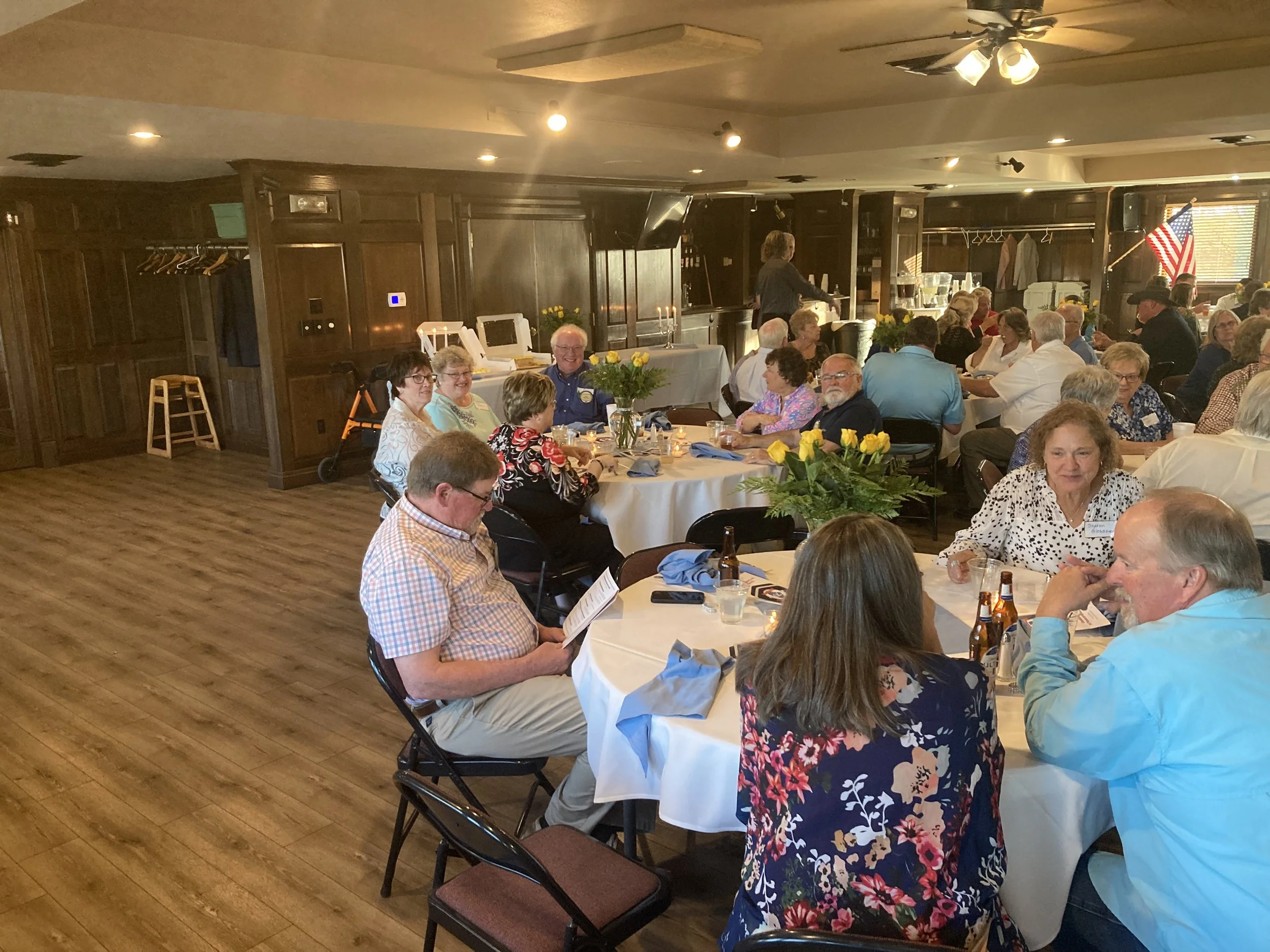 A group of people gathered at round tables in a banquet hall, dining and socializing. The room has dark wood paneling, a wooden floor, and is decorated with yellow flowers. An American flag is visible in the background, along with a stage and a perso
