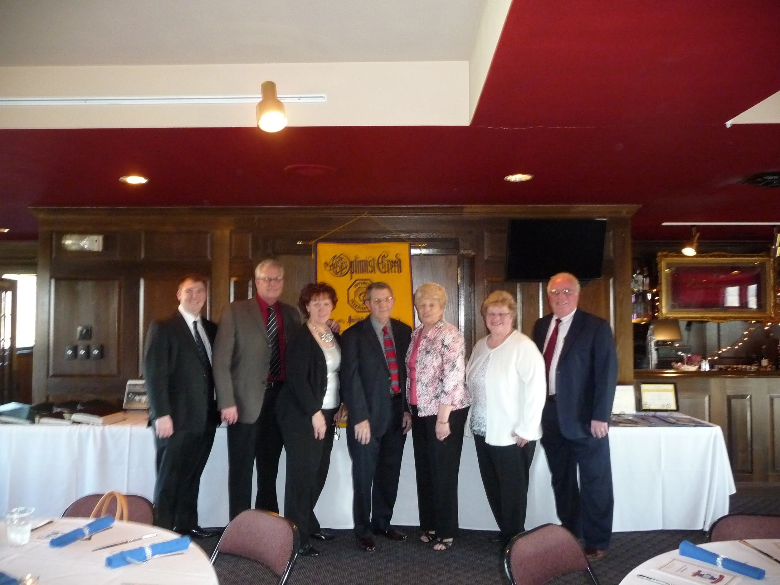 Group of seven people standing in a restaurant or banquet room at the Waterloo Optimist Club anniversary celebration, dressed in formal or business attire, with a yellow banner and a TV behind them.