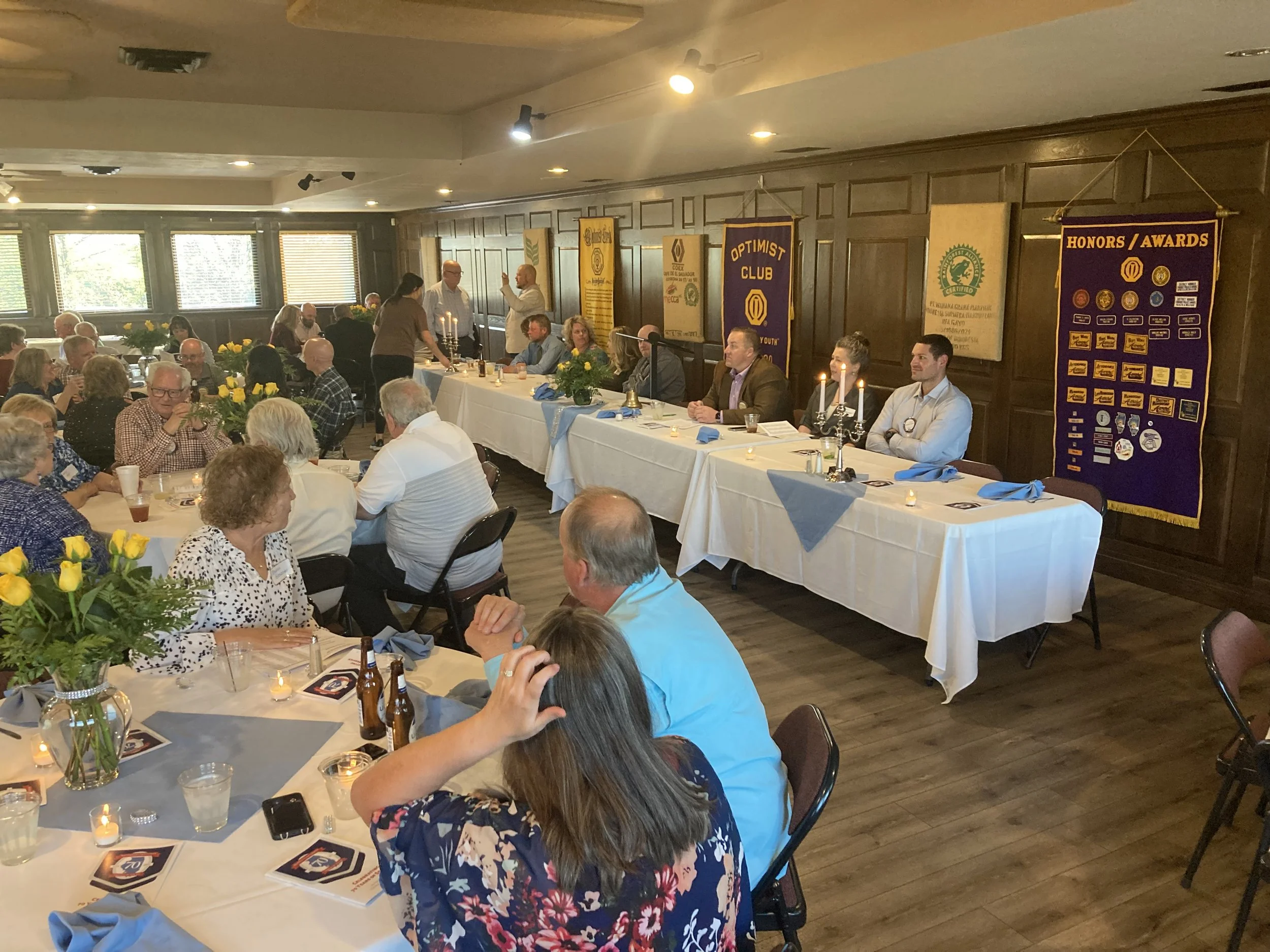 A group of people seated at round tables in a decorated room, attending a meeting or celebration, with banners on the wall, candles on the tables, and some people speaking at a long head table.