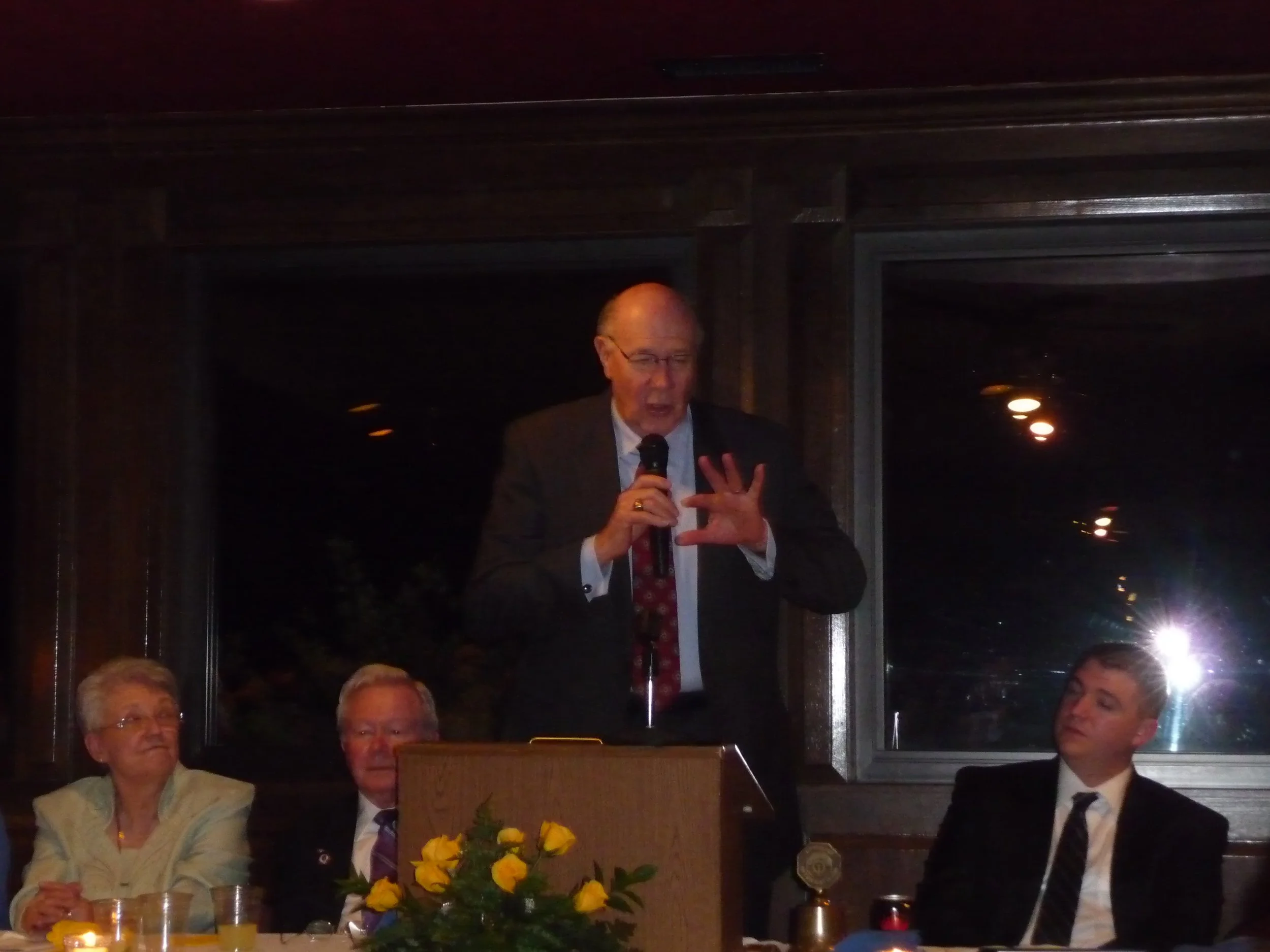A man in a suit and tie speaking at a podium with a microphone in a dimly lit room, seated at a table with three others, a woman and two men, with a bouquet of yellow roses in front of him. The woman and one man are looking at him, while the other ma
