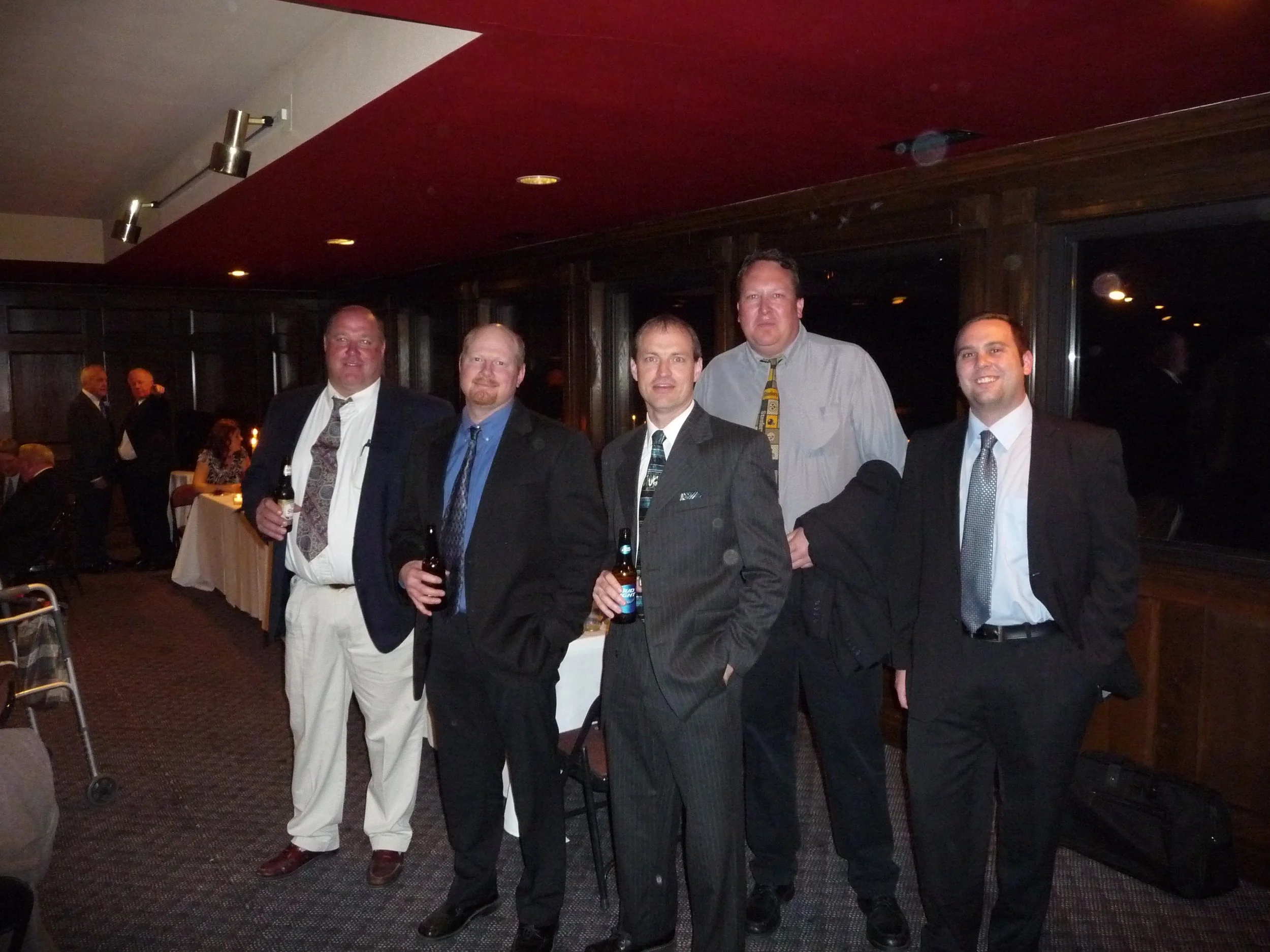 Six men in suits and one in a dress shirt with a tie, standing together indoors at a social gathering, some holding drinks, in front of a dark wooden wall with windows and men in the background.