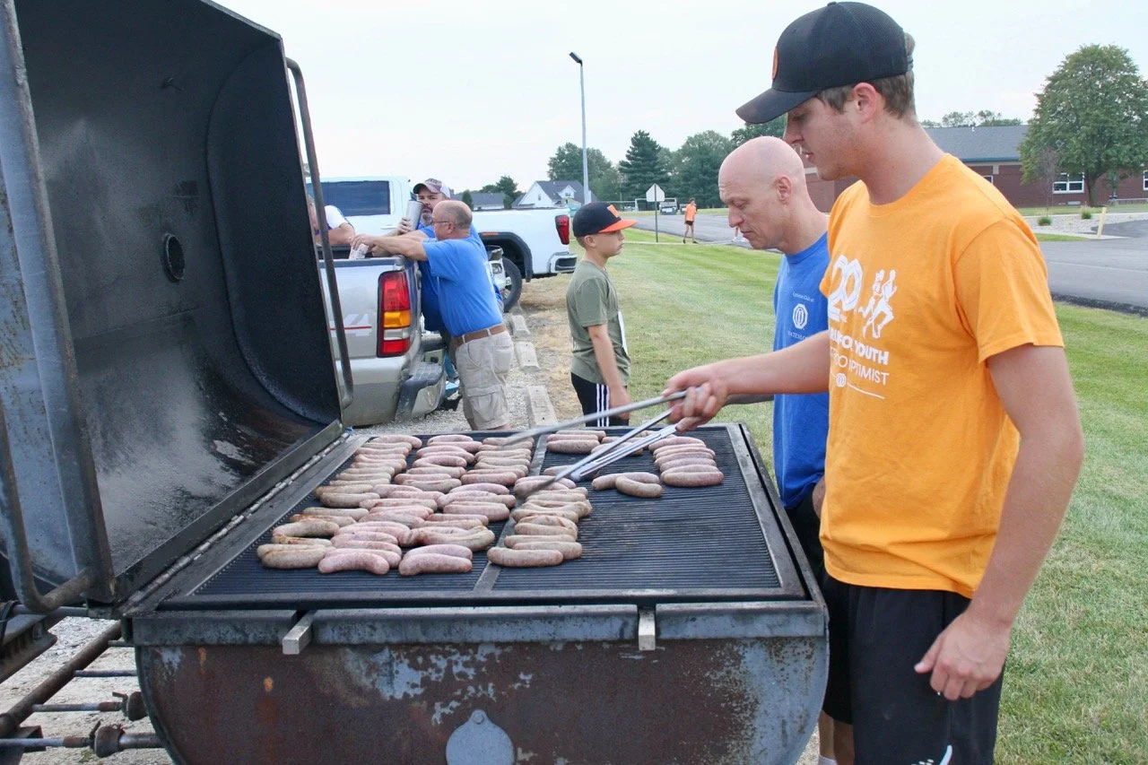 Waterloo Optimist Club members grilling sausages on an outdoor barbecue grill, with other people standing nearby for a community event.