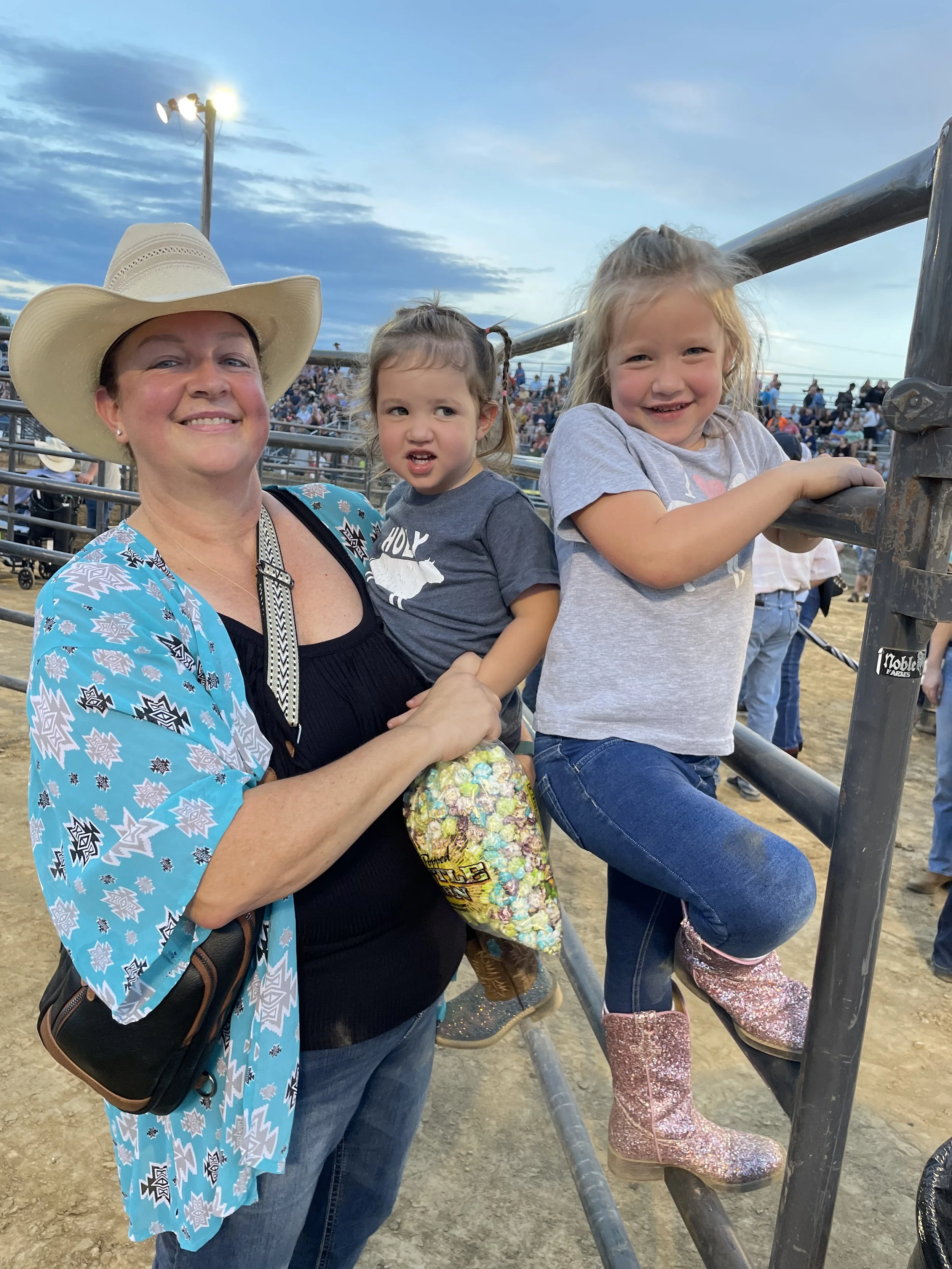 A woman wearing a cowboy hat and blue patterned shirt holding two young girls at the Blue Army Bull Bash. The girls are sitting on a metal fence, one with sparkle boots and the other with messy hair. 