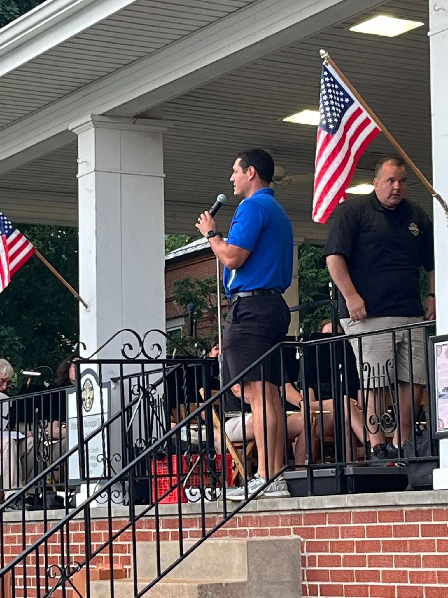Optimist Club member speaking into a microphone on a stage with American flags, standing next to a security guard.