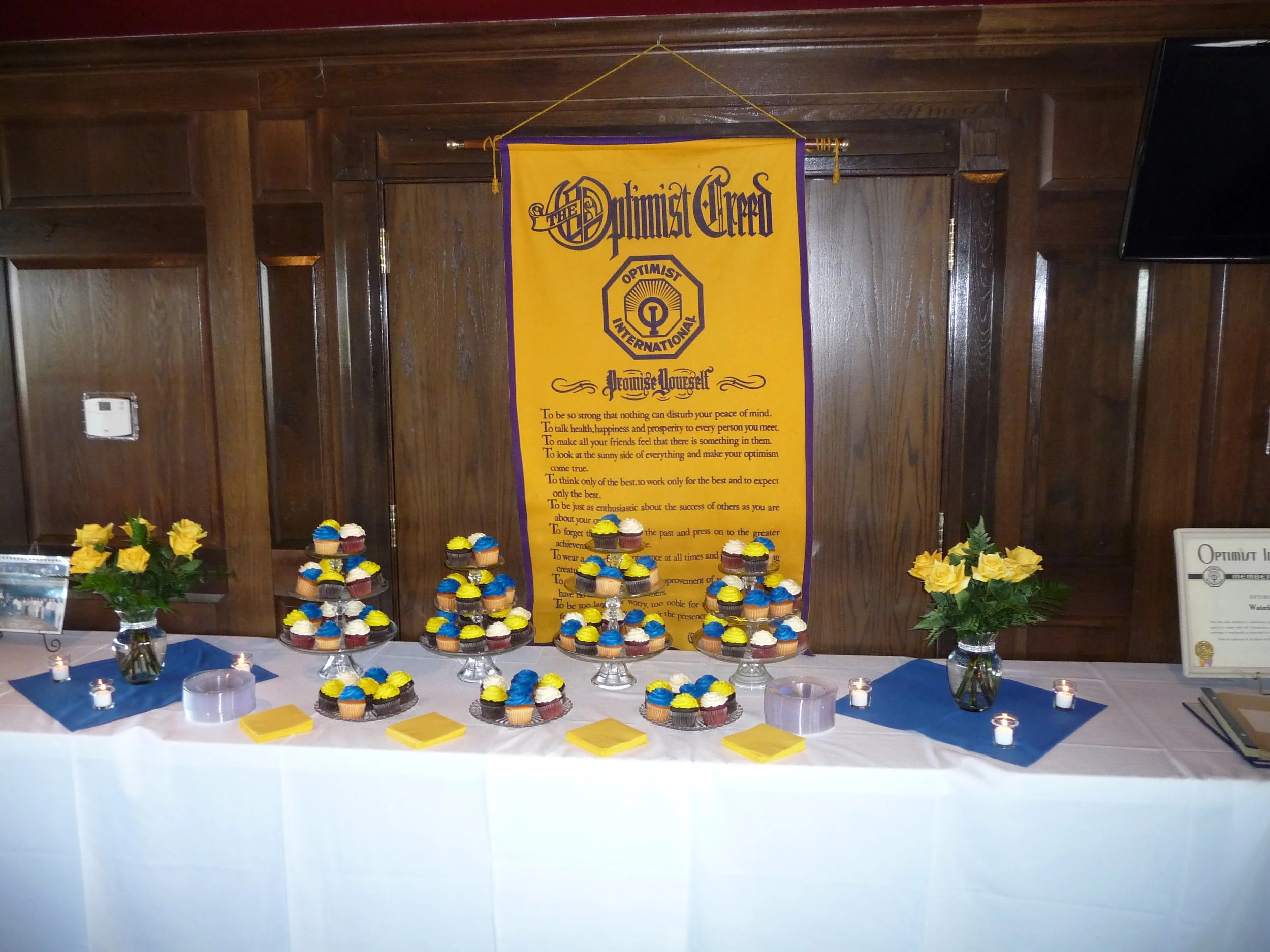 A table with cupcakes decorated in yellow, blue, and white, yellow roses in vases, yellow and blue napkins, and small candles, against a wooden wall with a yellow Optimist Club banner.