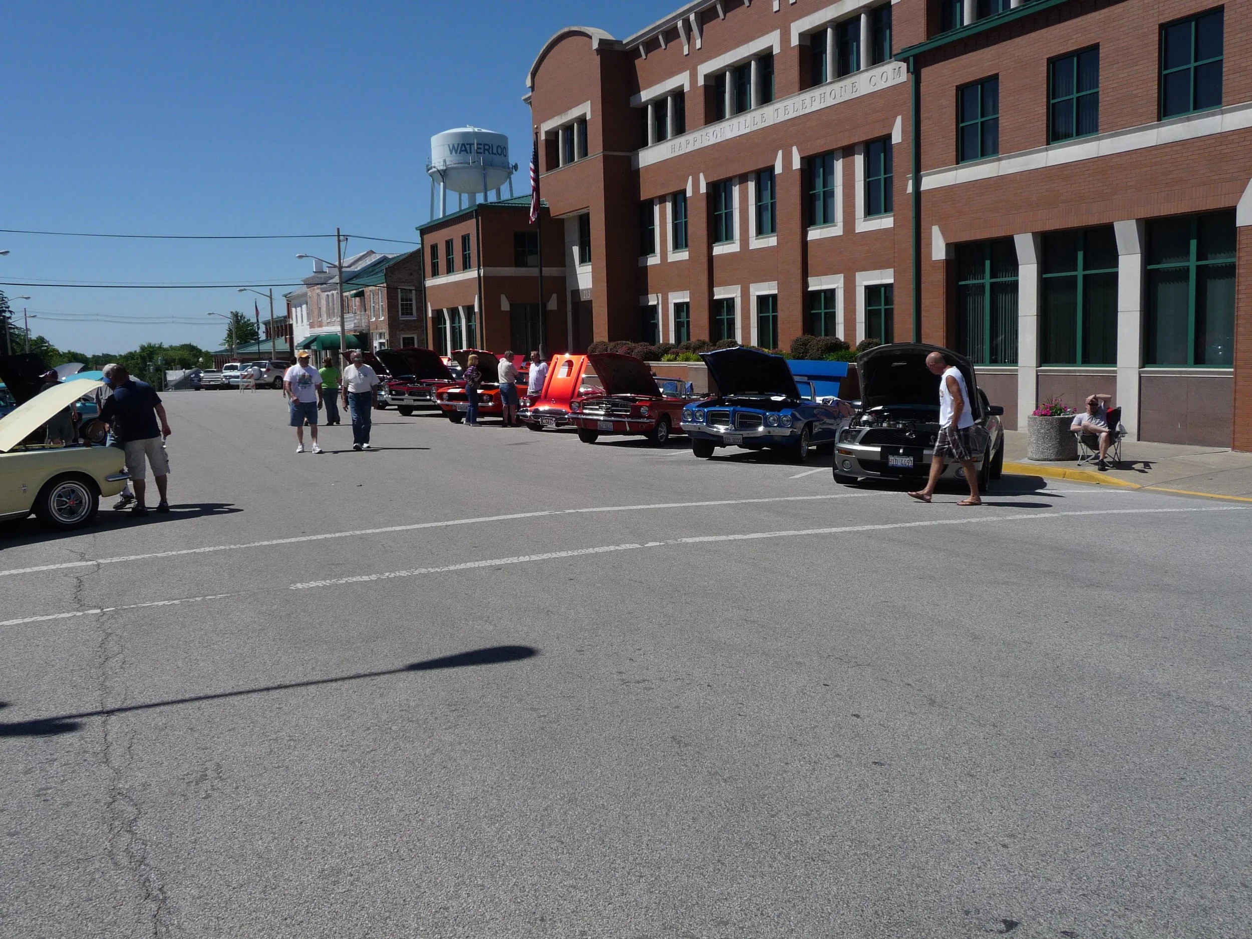Lineup of vintage and classic cars with open hoods parked along a city street during the Waterloo Optimist Club car show, with people walking and observing the vehicles.