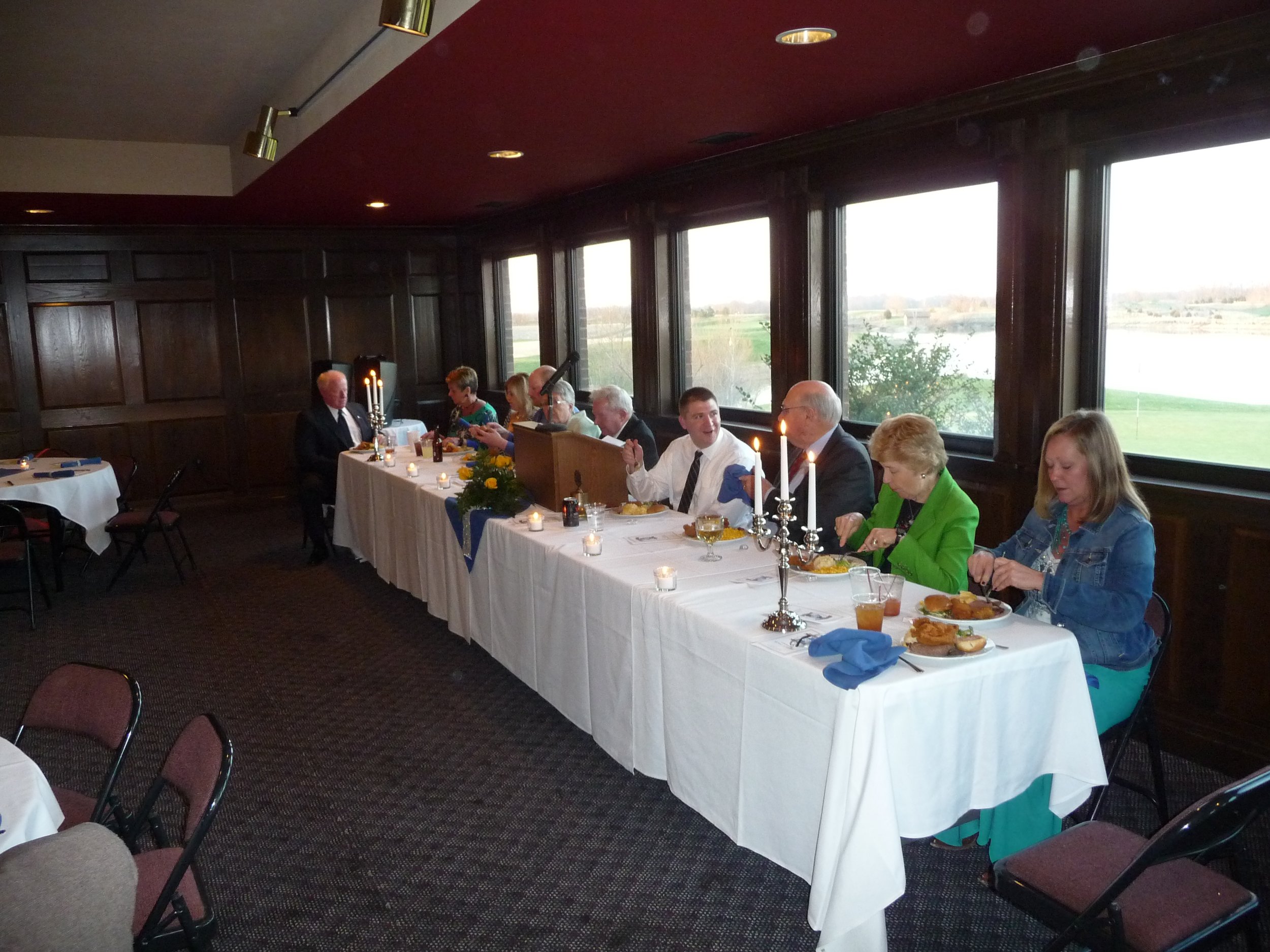 People sitting at a long banquet table with white tablecloths, candles, and centerpieces, dining indoors near large windows showing a scenic outdoor view at the  Waterloo Optimist Club anniversary celebration. .