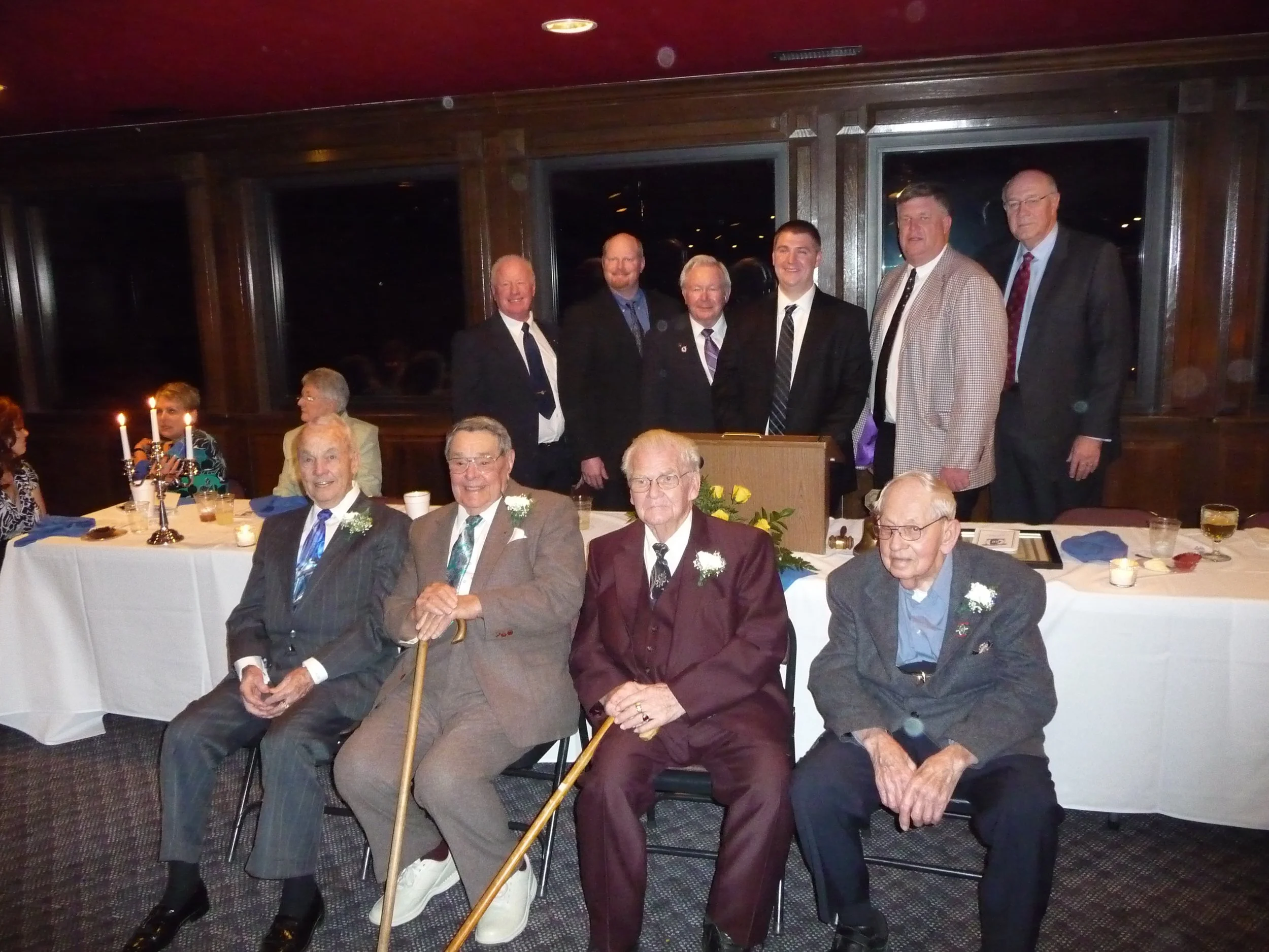 Group of elderly men and middle-aged men at a formal gathering or celebration, some wearing suits with flower boutonnieres, seated and standing in a room with wood paneling and white tablecloths on tables.