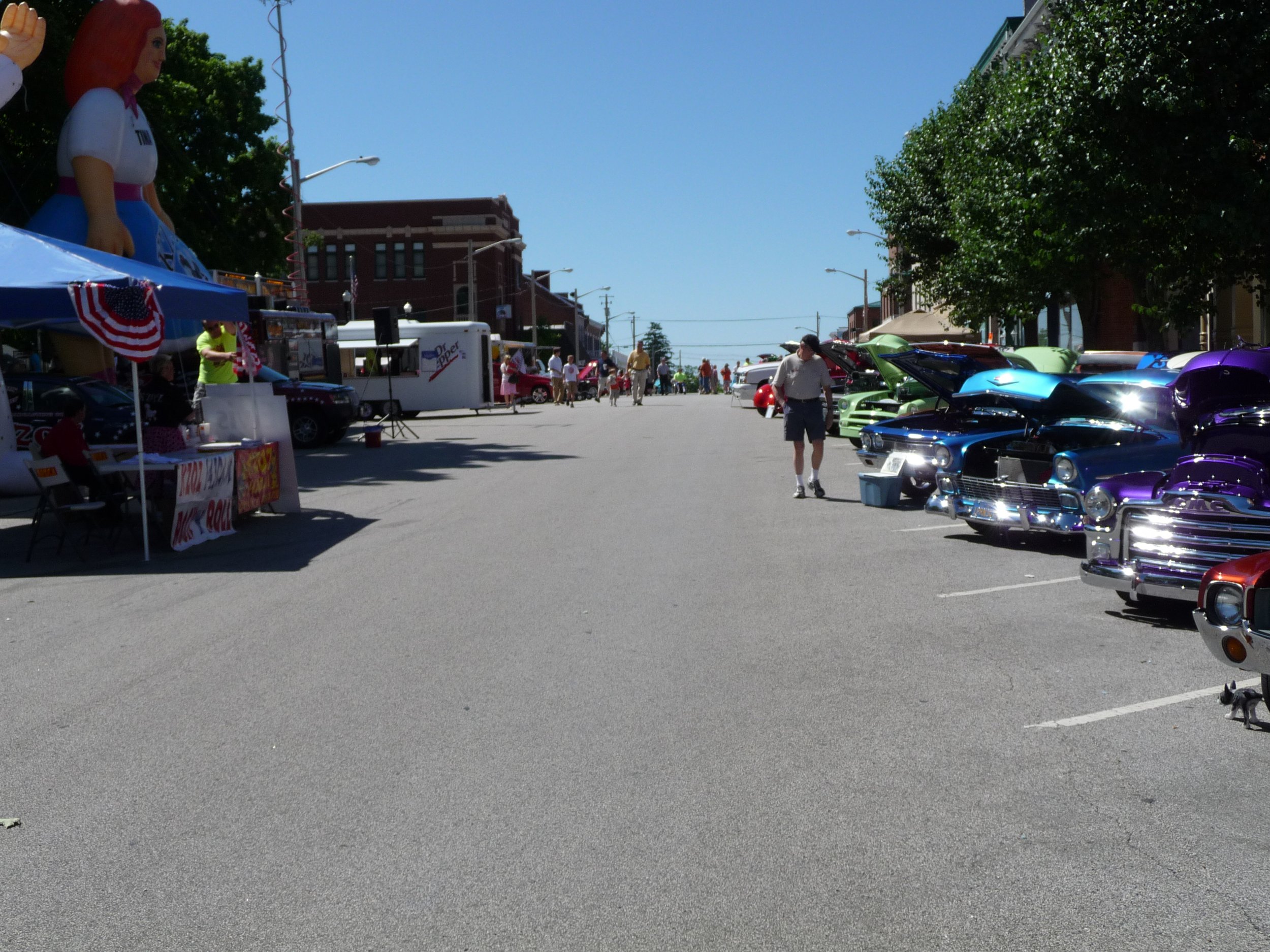 Waterloo Optimist Club car show with vintage cars parked along the right side, vendor stalls on the left, and a large inflatable doll visible on the left. People are walking and browsing the event on a clear, sunny day.