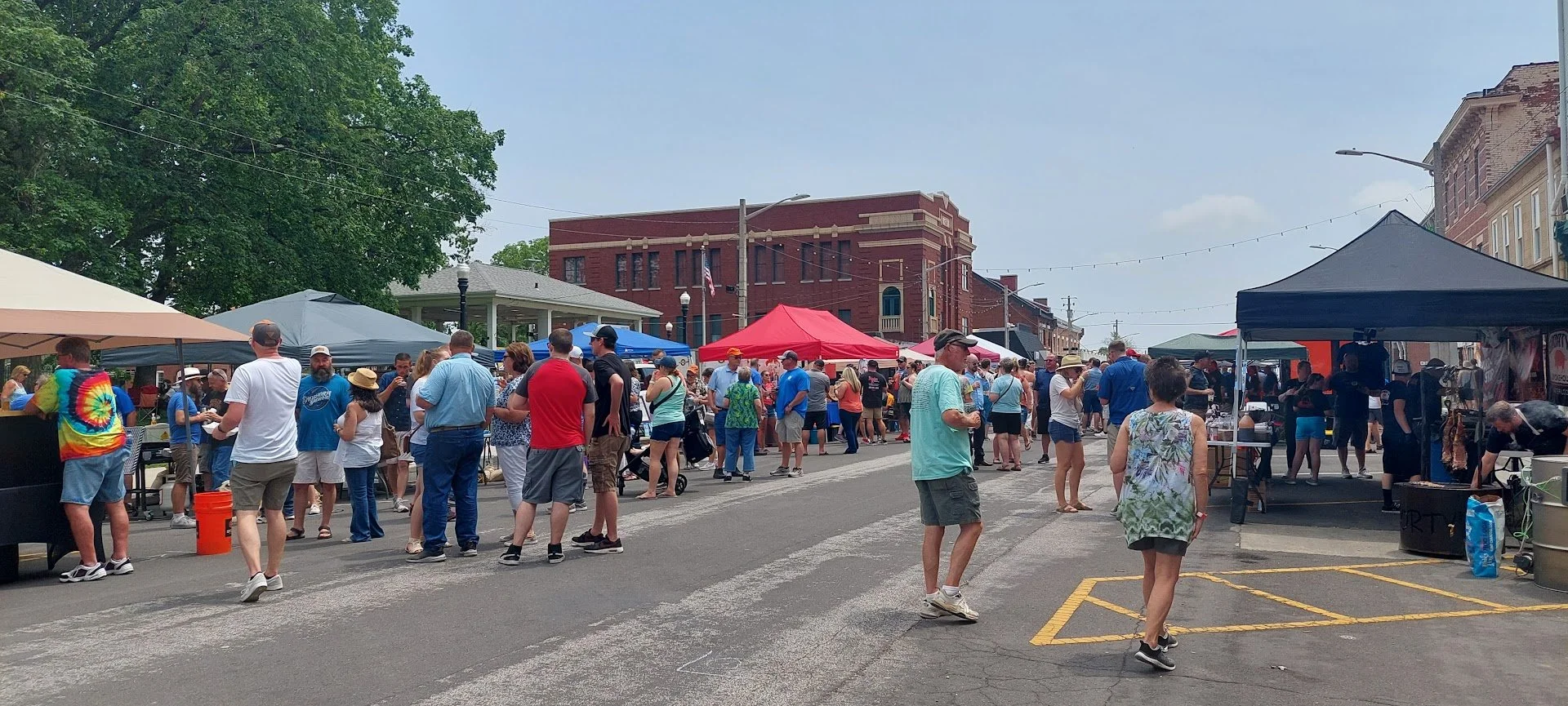 People walking and browsing at the Beer-B-Que with vendor tents and booths on a city street under a partly cloudy sky.